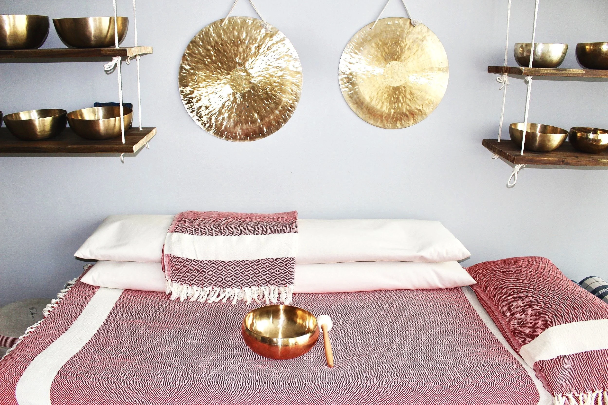 Bett mit weißem Kopfkissen, einer roten Decke und einem goldenen Klangschale auf den Decken. Über dem Bett an der Wand hängen zwei goldene Klangplatten, in der Nähe sind Wandregale mit weiteren goldenen Klangschalen.