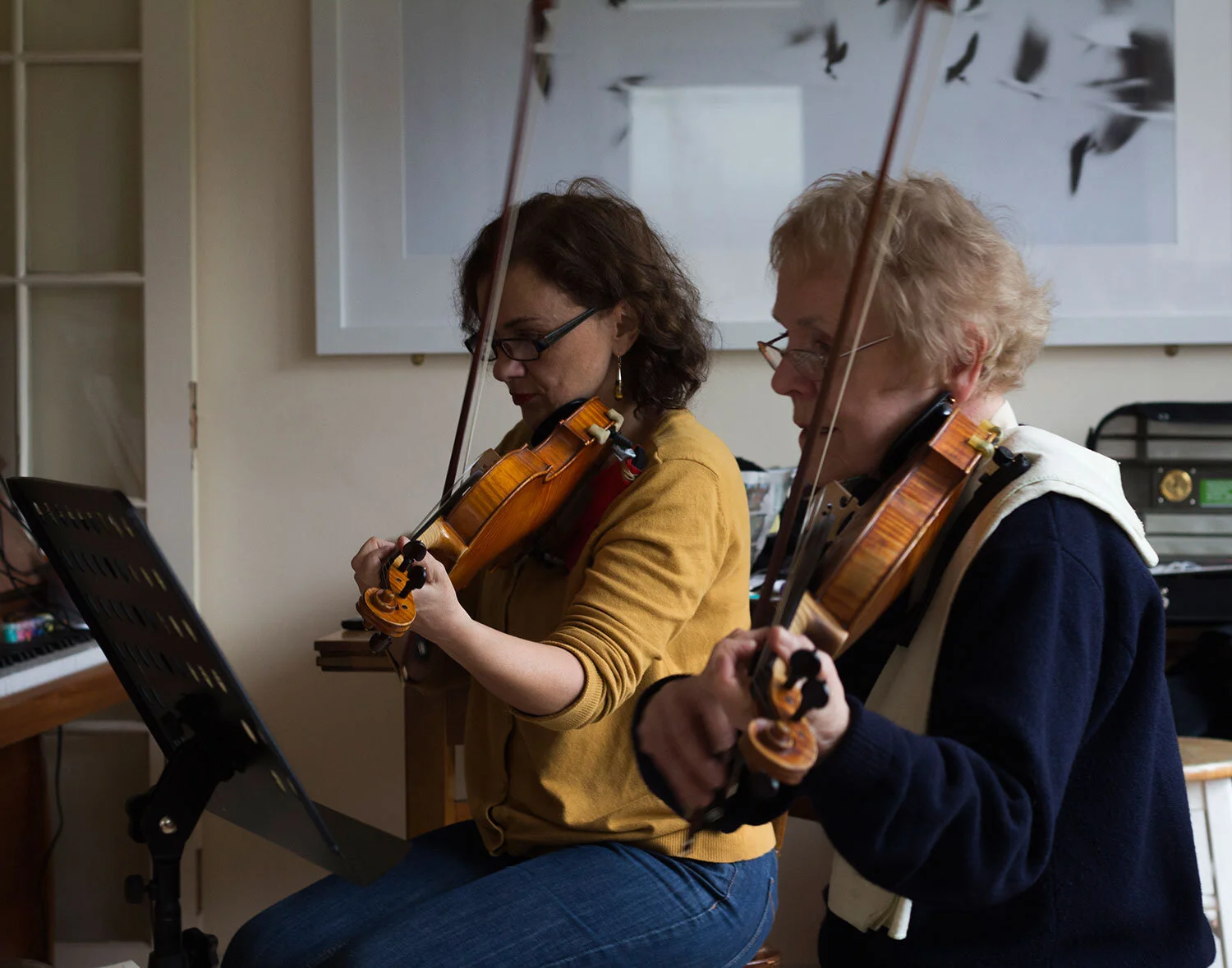 Two women playing violins in a room, seated side by side, reading music from a stand in front of them.