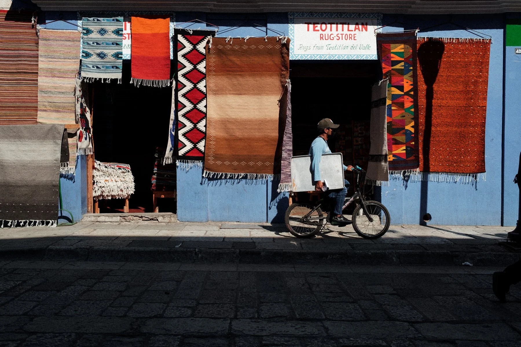 Oaxacan street photography: a cyclist passing by a traditional rug shop, captured by Lucca Lazzarini.
