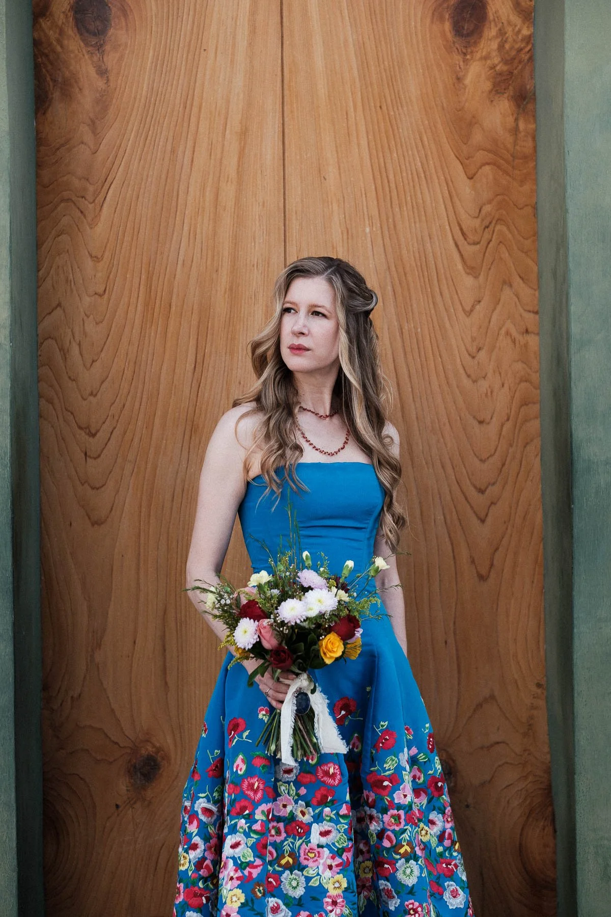 A documentary-style portrait of the bride in her blue dress holding a colorful floral bouquet.
