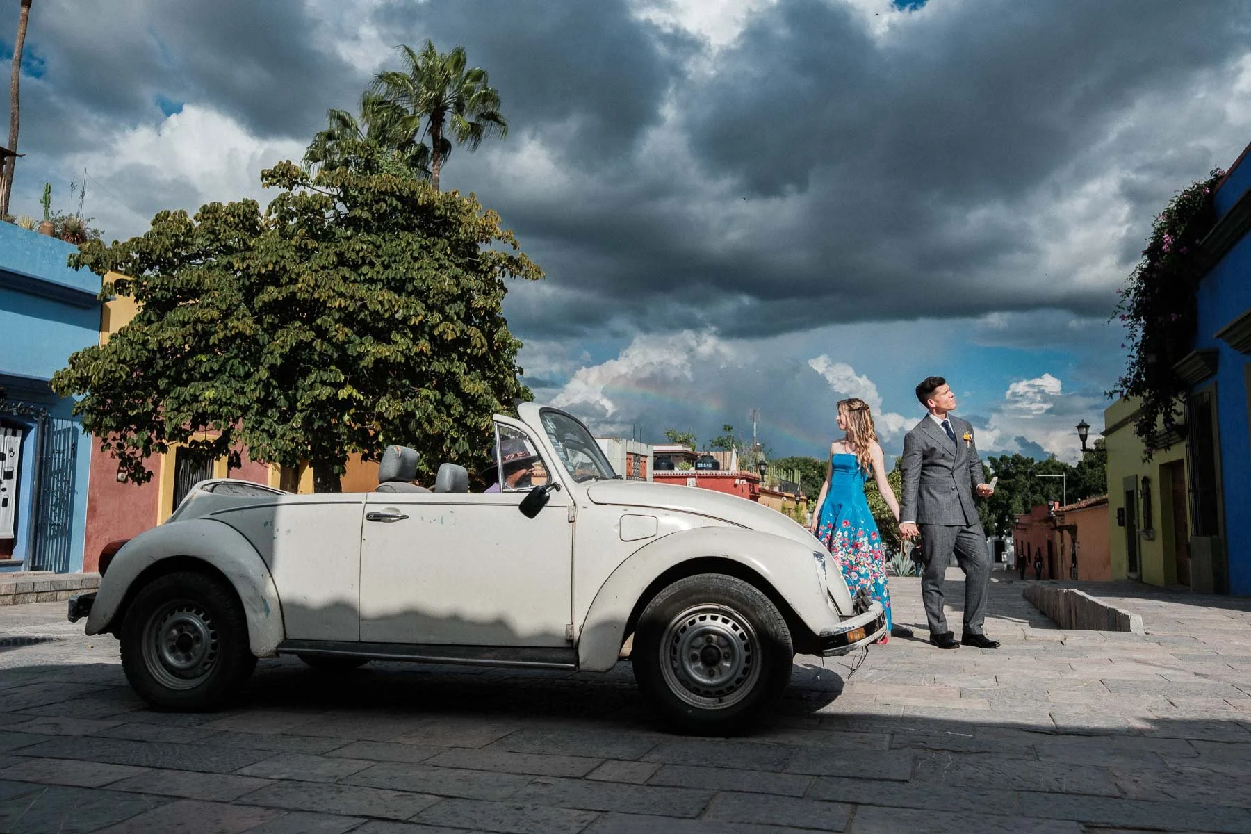 A classic "Vocho" passing under a rainbow and storm clouds during an unscripted wedding moment.