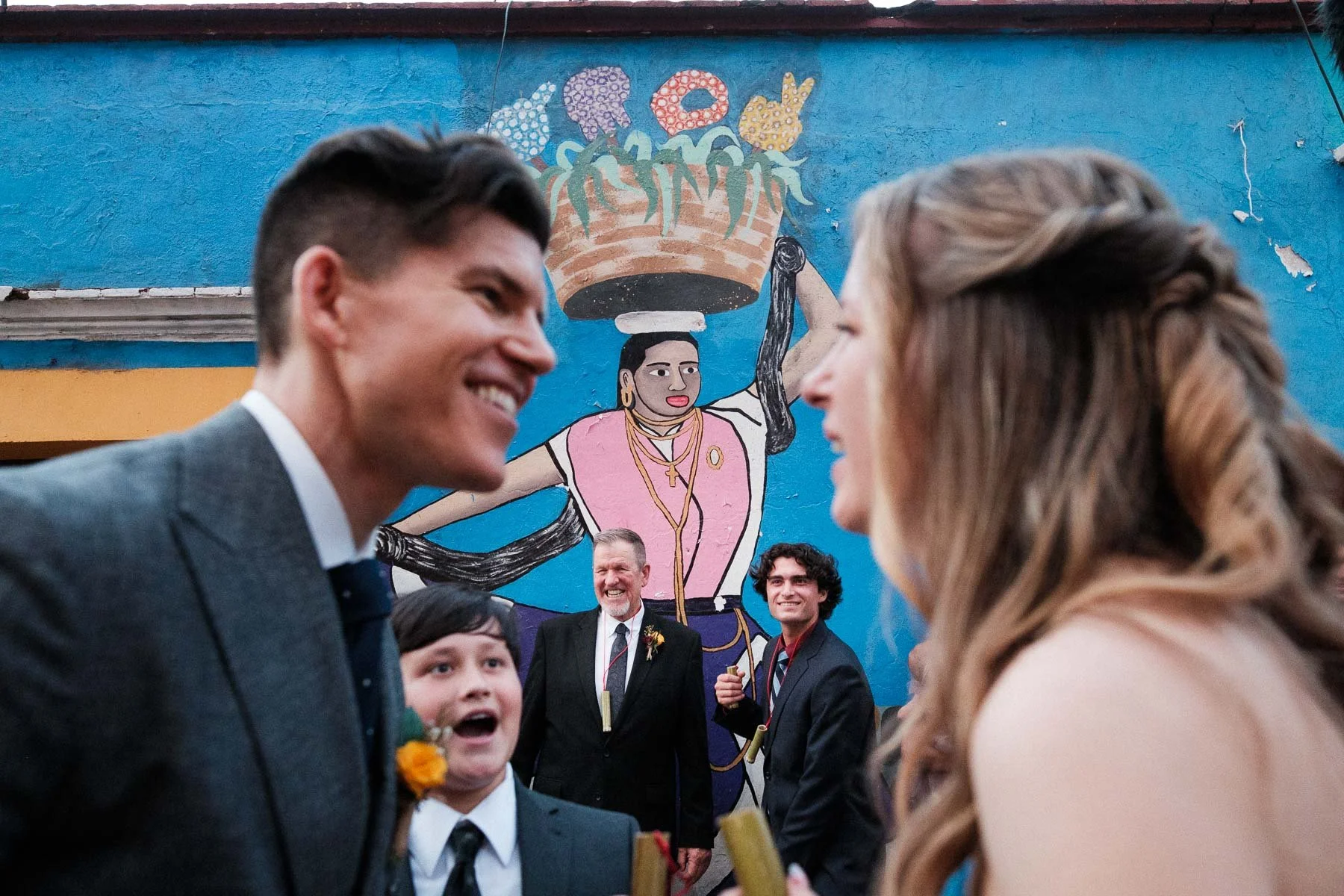A complex, layered frame of the couple looking at each other, framed by a dancer mural and family in the distance.