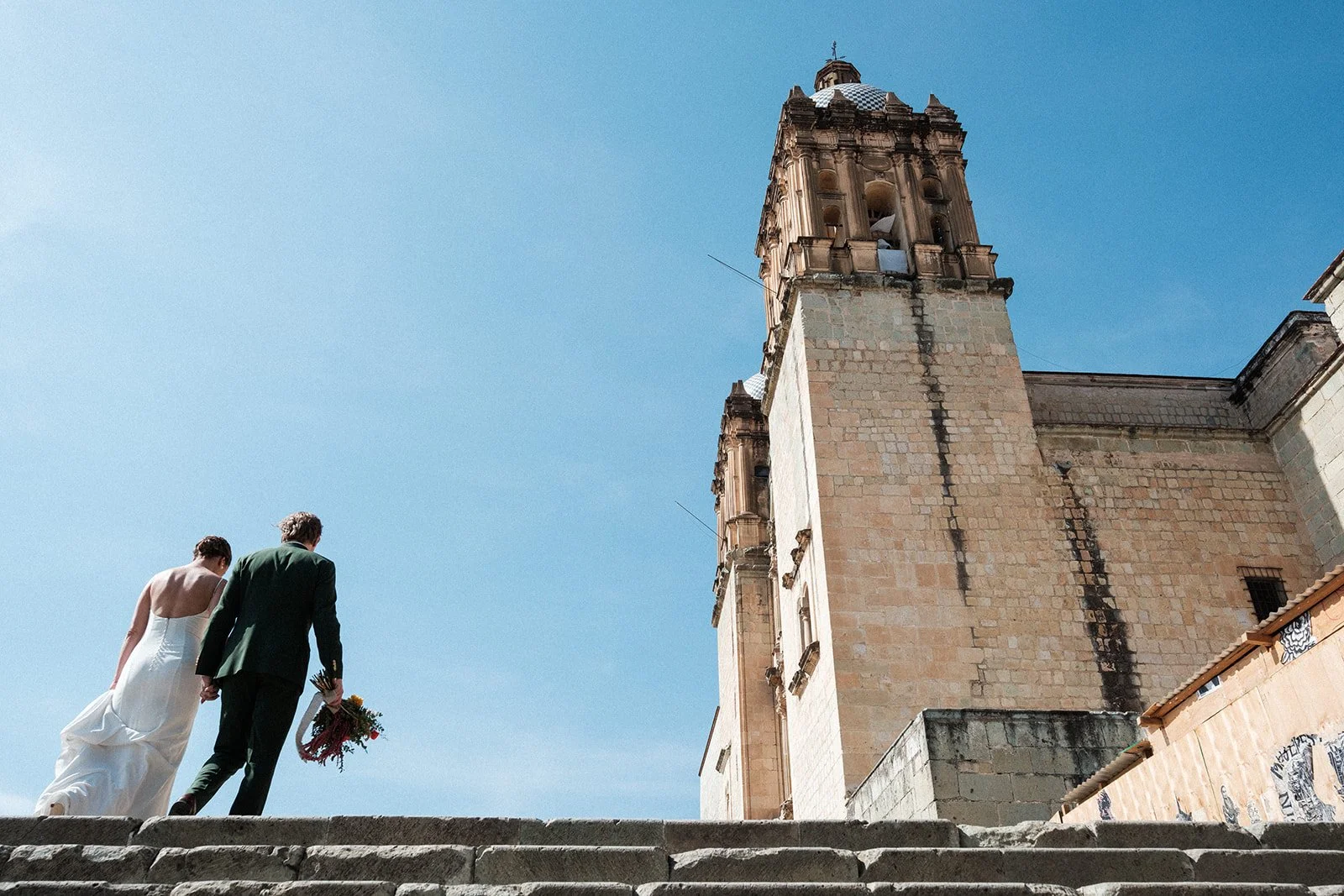 Bride and groom walking up stairs with the iconic Santo Domingo Church and a blue sky in Oaxaca.