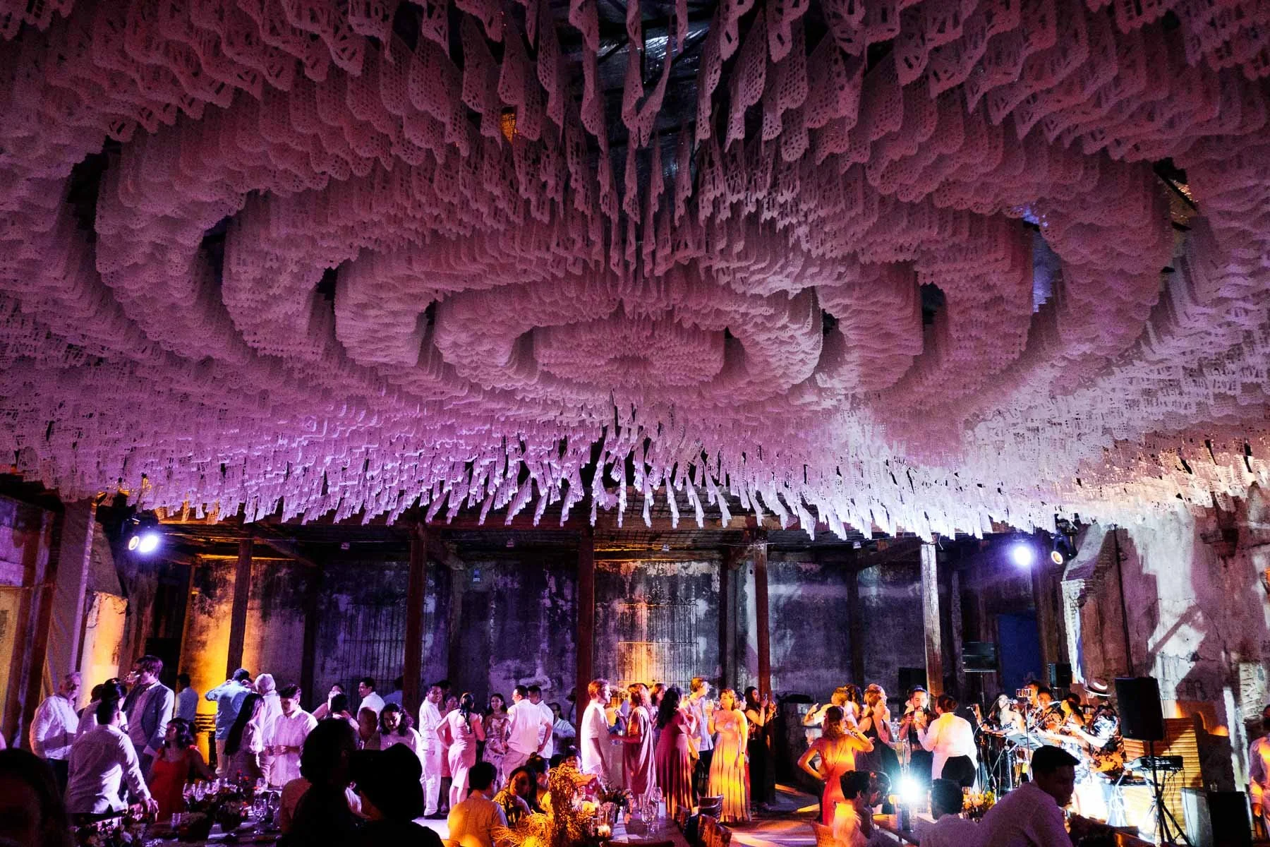 Nighttime wedding celebration at Proyecto Murguia in Oaxaca featuring colorful Mexican Papel Picado banner details and dramatic violet light on historic ruin walls.