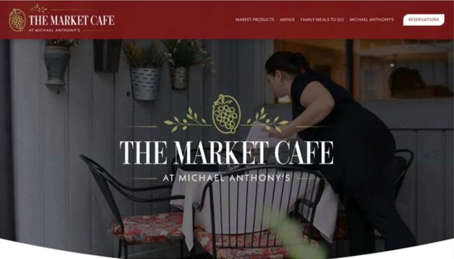 Website homepage for The Market Cafe at Michael Anthony's, showing a woman setting a table outside on a patio with potted plants and a gray wooden fence.
