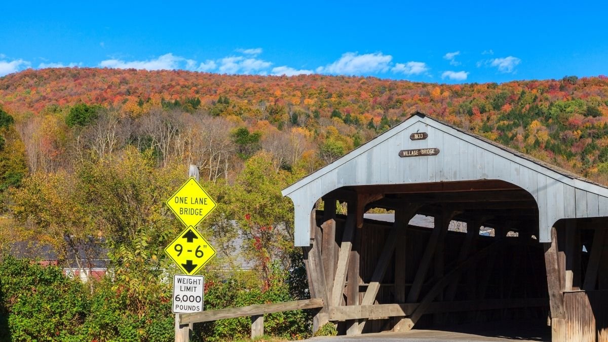 Historic Waitsfield Covered Bridge spanning a valley, with a yellow road sign showing a weight limit and arrow beneath a blue sky and autumn-colored hills.