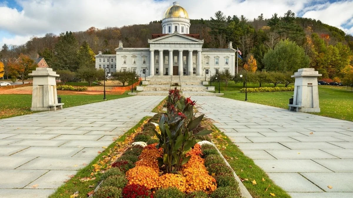 Vermont State House in Montpelier; a white-domed neoclassical building with columns, viewed from a stone walkway lined with autumn trees and a flowerbed