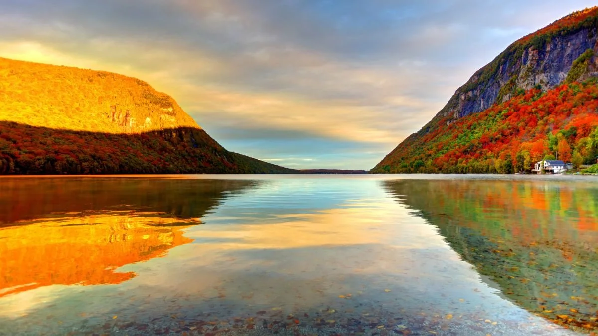 Calm sunset at Lake Willoughby, VT, with mirrored reflections, a rocky hillside on the left glowing orange, and a green-and-red autumn hillside on the right.