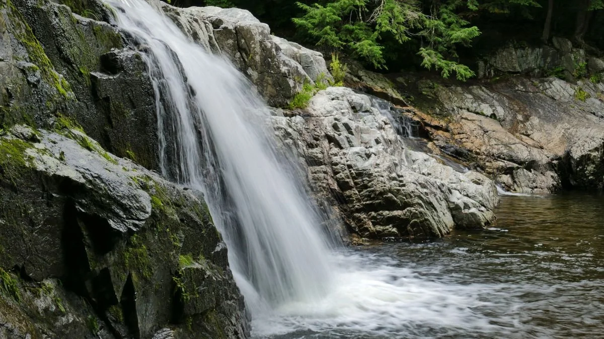 A wide cascade of water pours over dark rocky cliffs into a calm river at Buttermilk Falls, Ludlow, Vermont, surrounded by moss-covered rocks and lush trees.