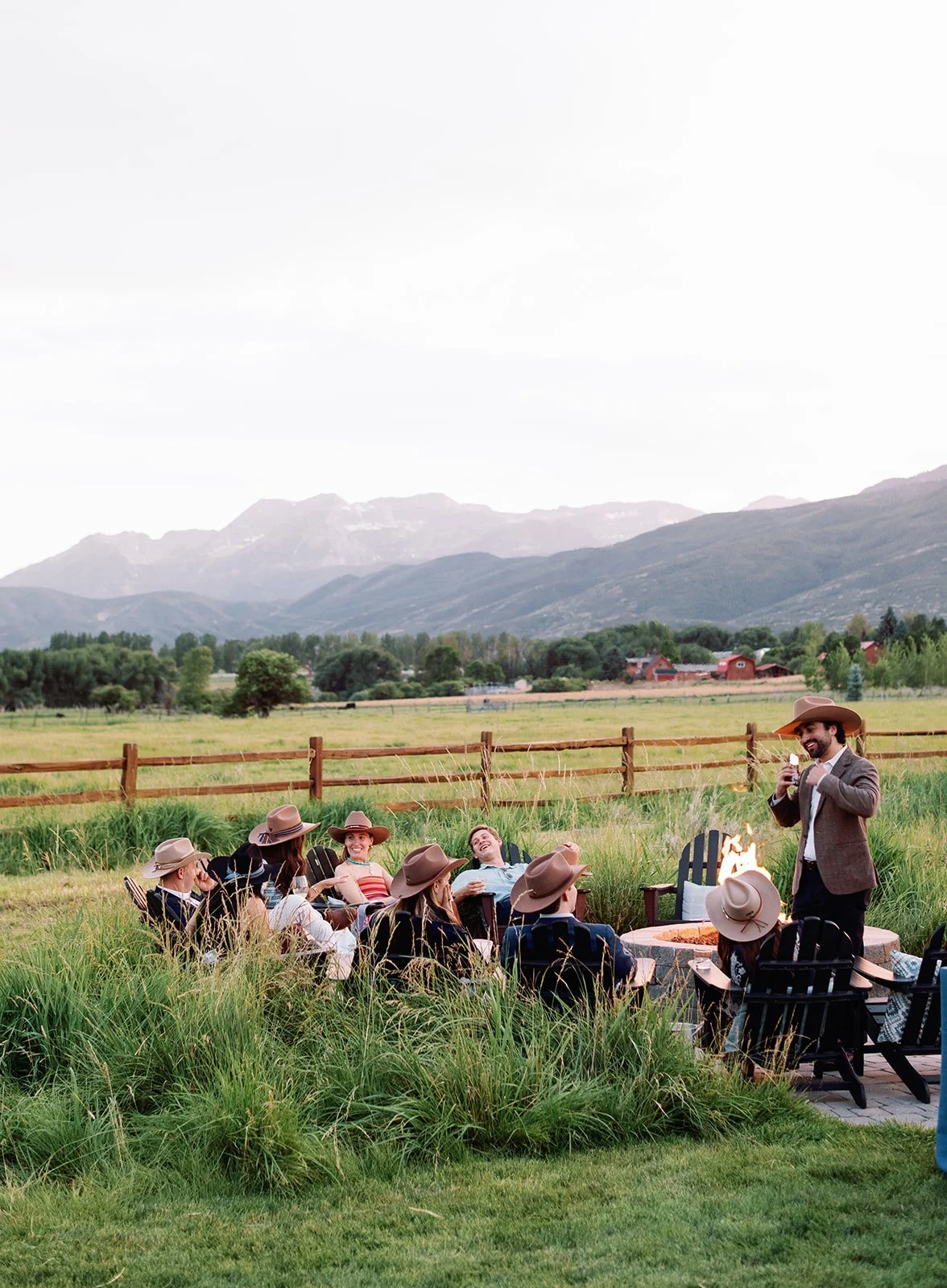 Wedding guests wearing customized cowboy hats relax around an outdoor firepit with sweeping mountain views at River Bottoms Ranch in Utah. This Western-chic gathering was part of a luxury destination wedding weekend planned and designed by Michelle L