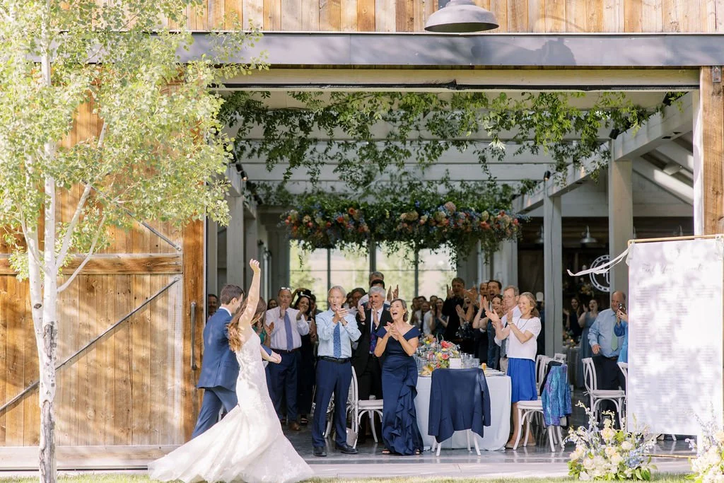 Newlyweds make a joyful entrance into the barn during a wedding dinner reception at 4U Ranch in Utah, surrounded by guests, floral installations, and rustic barn architecture designed and planned by Michelle Leo Events.