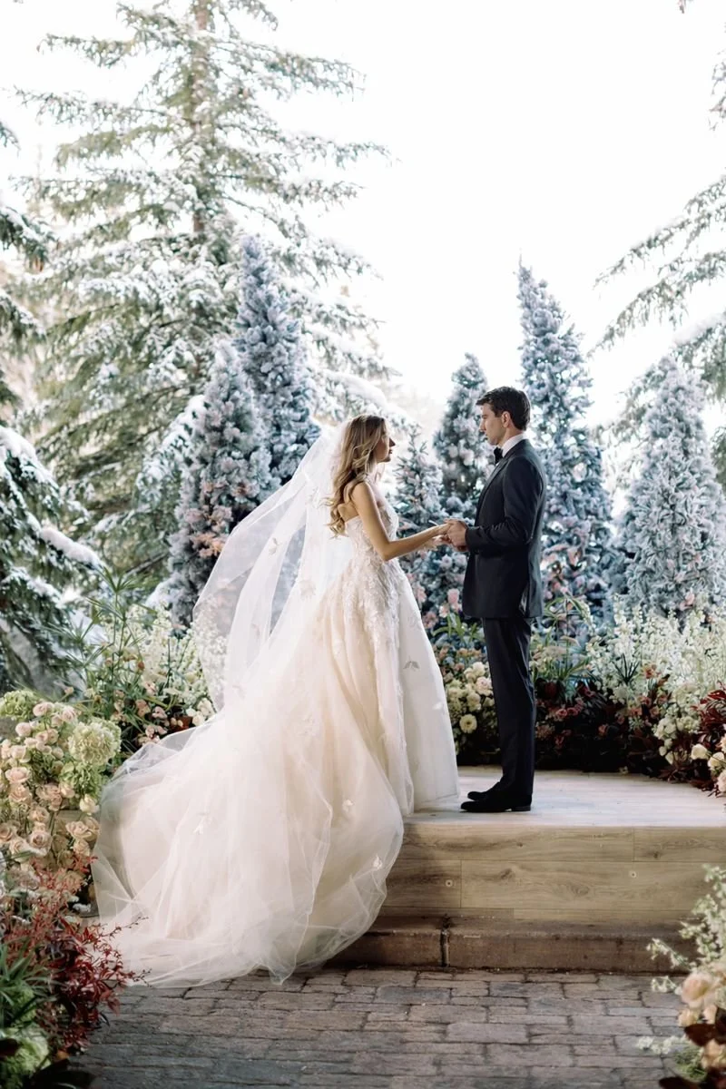 Catherine and Mitch exchanging vows during a winter wedding ceremony at St. Regis Deer Valley in Utah, surrounded by snow-covered evergreens and lush floral design.