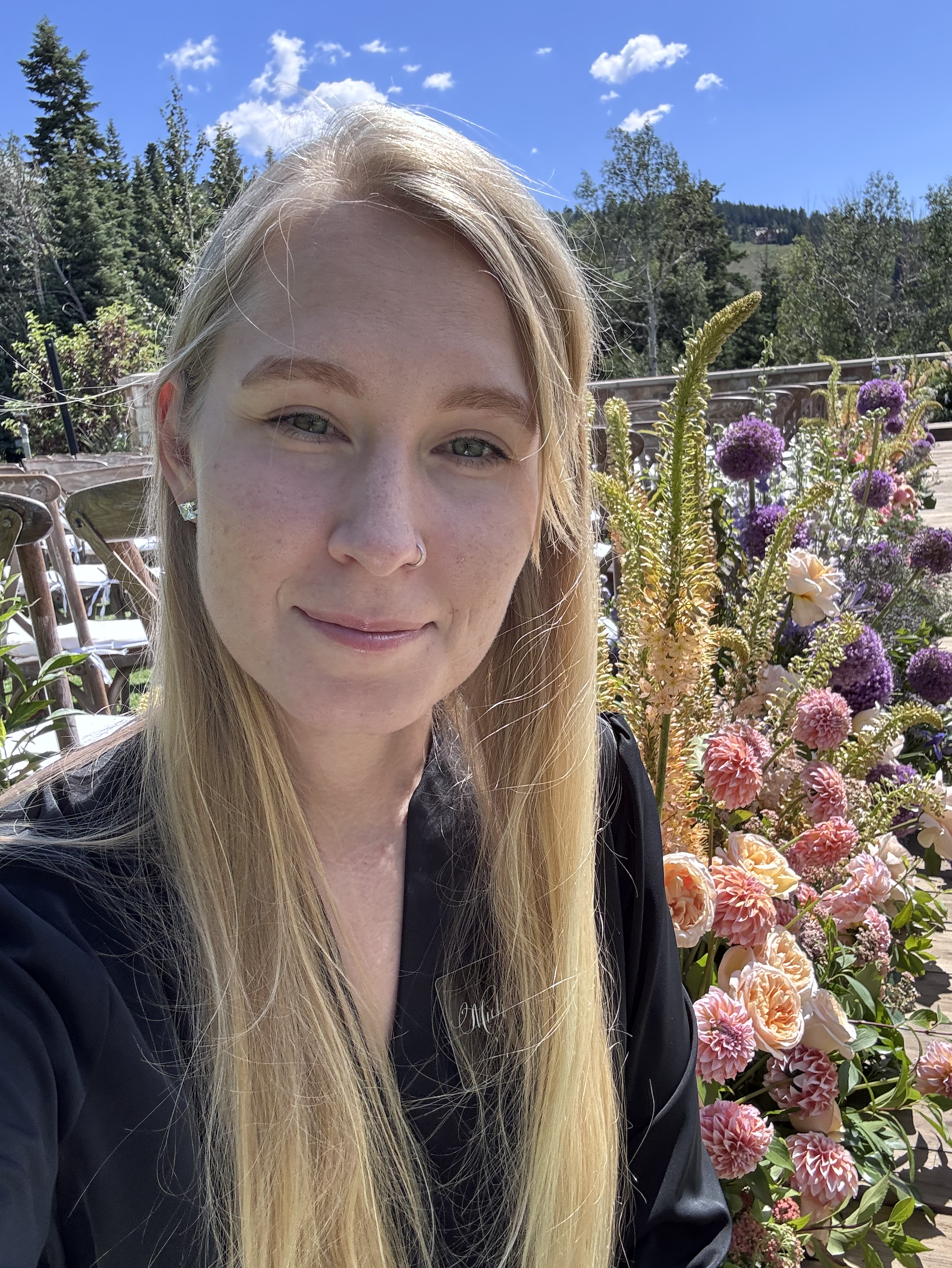 A young woman with long blonde hair, wearing earrings and a black jacket, taking a selfie outdoors next to pink and purple flowers. There are trees, a mountain, and a blue sky with scattered clouds in the background.