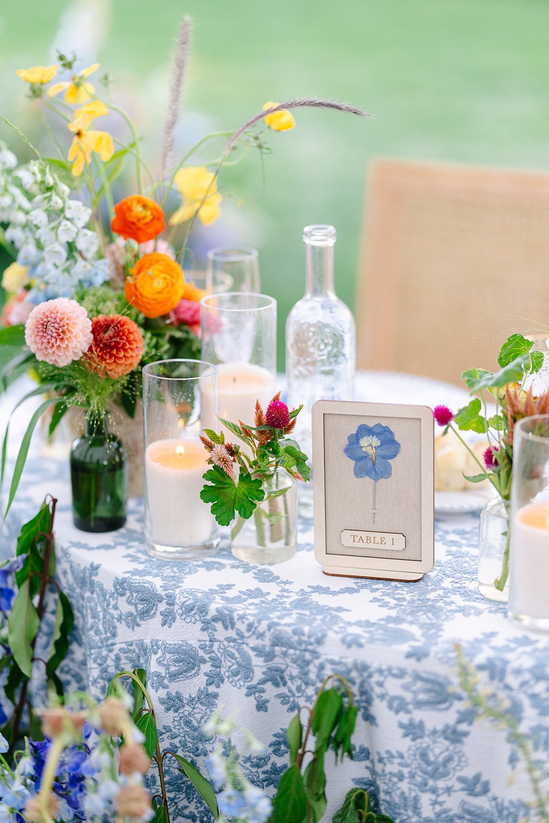 Colorful garden-inspired wedding centerpiece with orange and yellow flowers, blue patterned linens, candlelight, and personalized table number designed for a luxury Utah wedding by Michelle Leo Events.