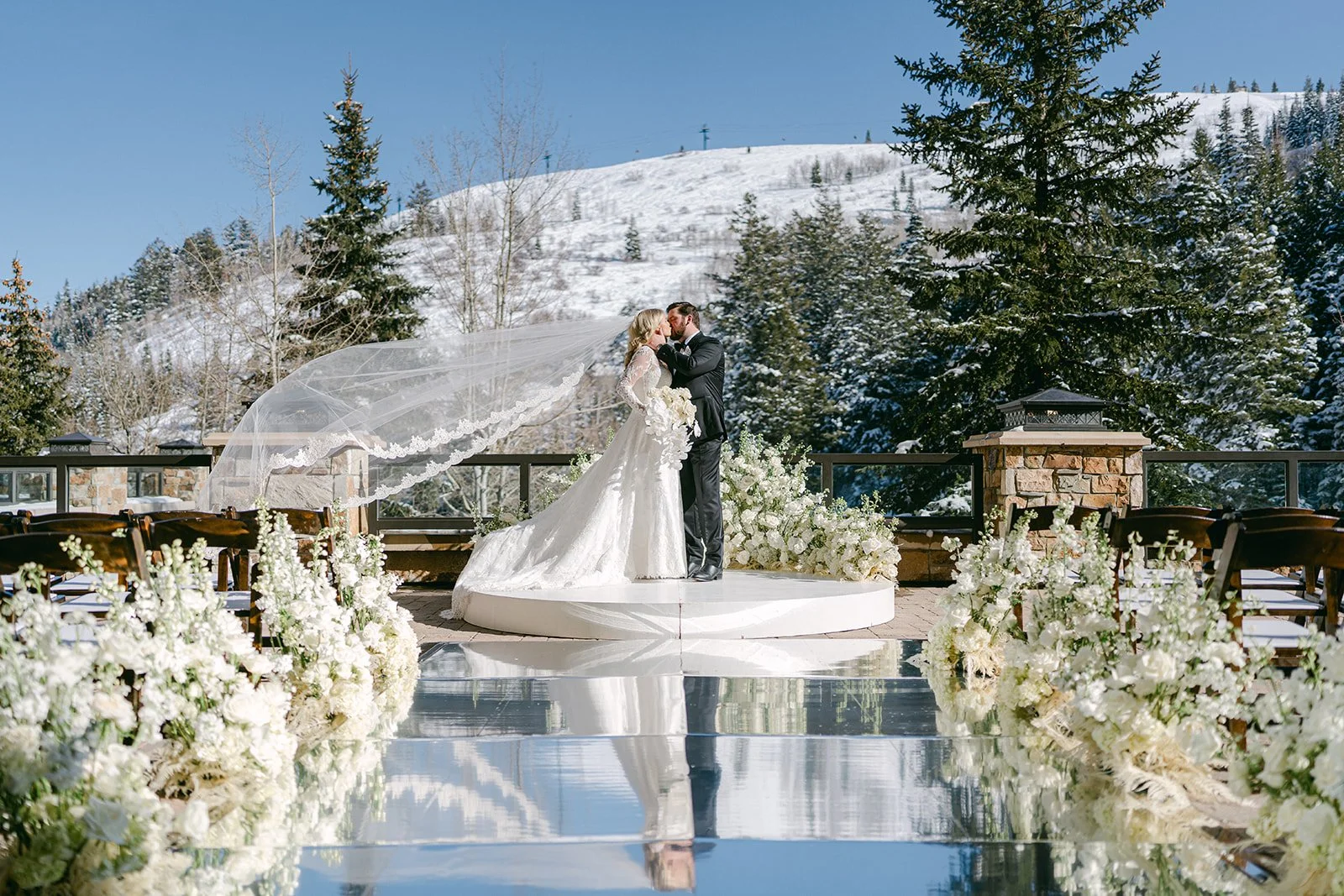 Bride and groom share a kiss during an outdoor winter wedding ceremony on the Astor Terrace at St. Regis Deer Valley, featuring a mirrored aisle, white florals, snow-covered mountains, and elegant ceremony