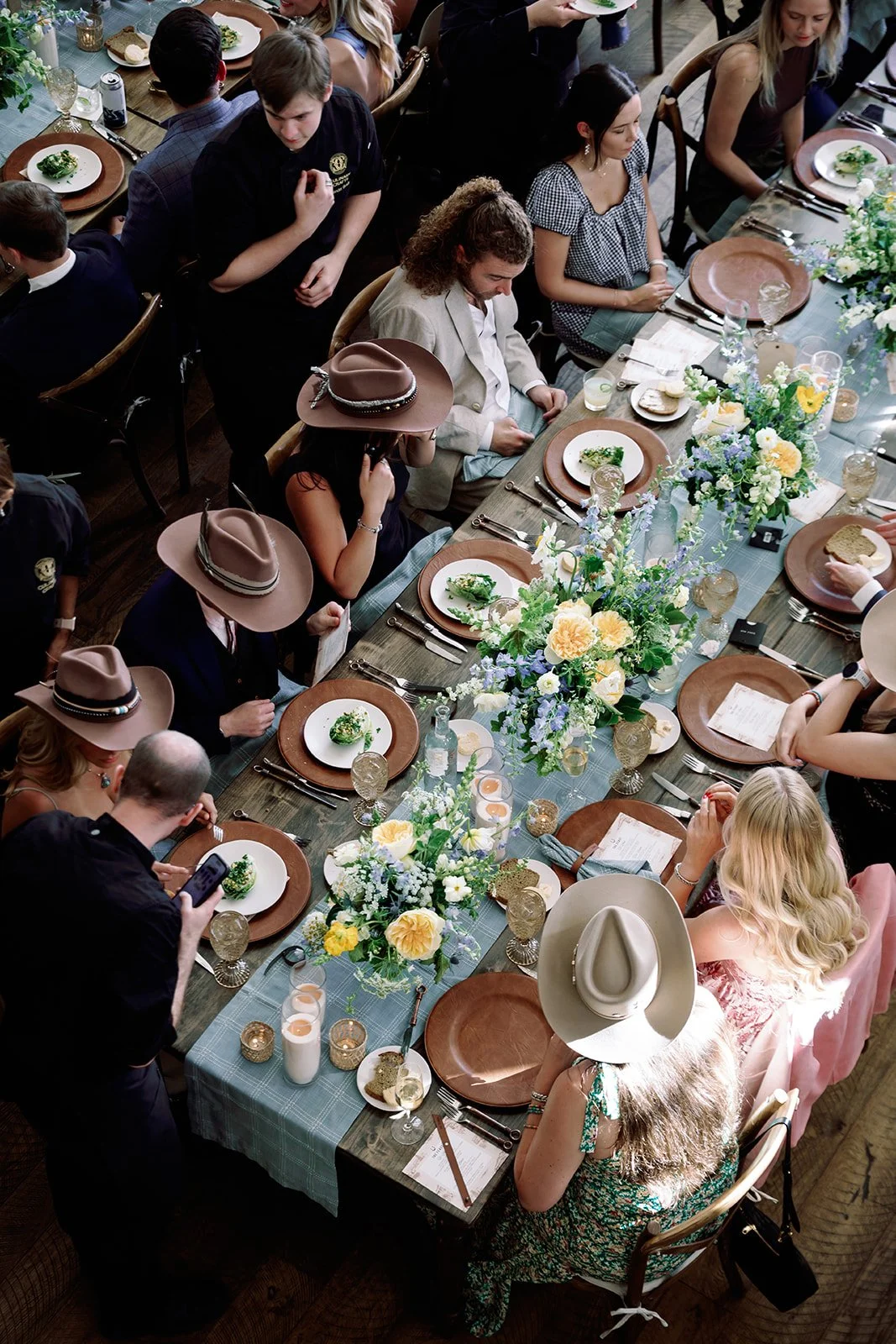 An overhead view of guests enjoying an elegant Western-chic rehearsal dinner at River Bottoms Ranch in Utah, wearing personalized cowboy hats around a beautifully designed table featuring blue linens, yellow and blue florals, and cognac chargers. Thi
