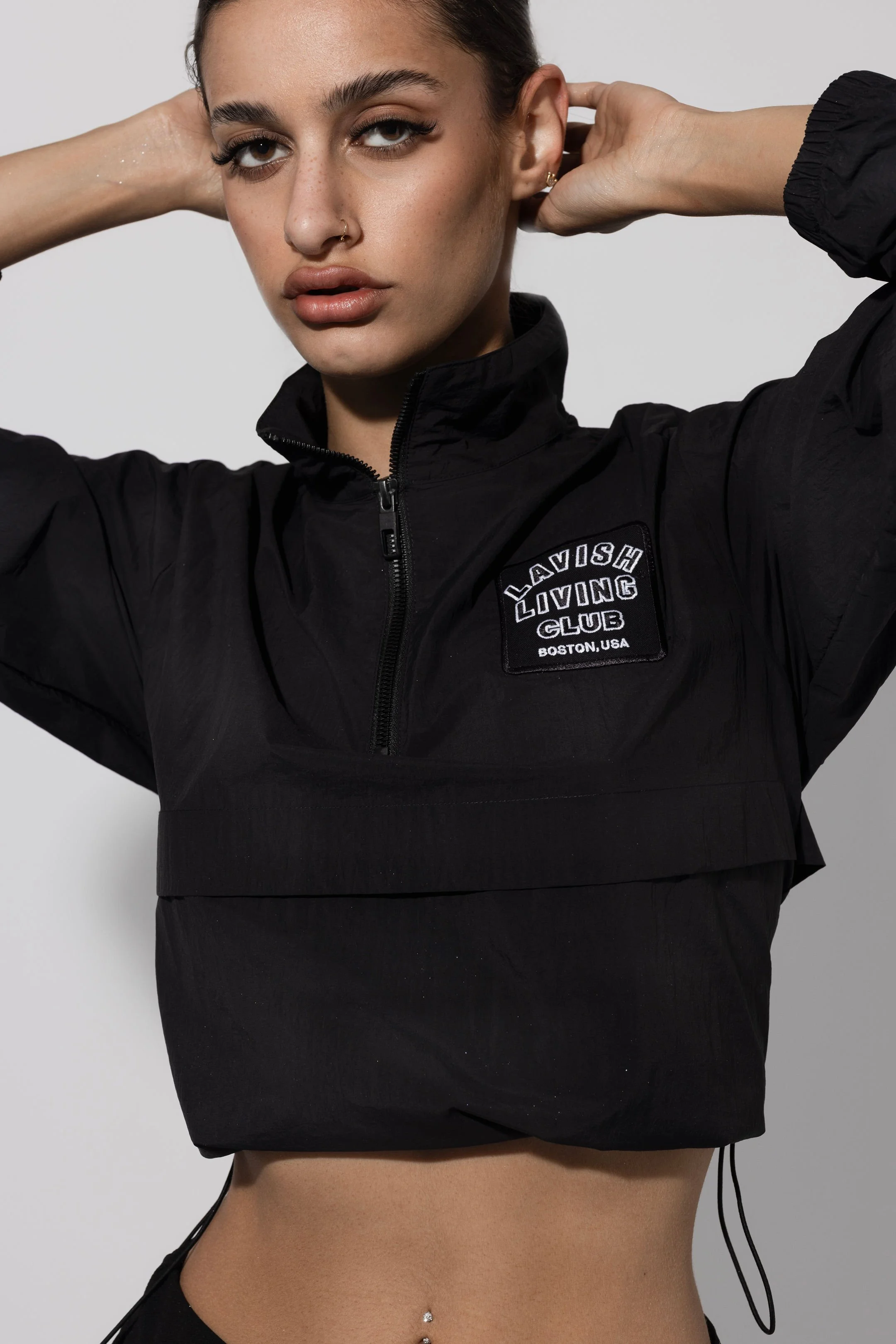 A woman with dark hair, nose piercing, and natural makeup poses with her hands behind her head, wearing a black black jacket with a patch that reads 'Lavish Loving Club Boston, USA' over a bare midriff.