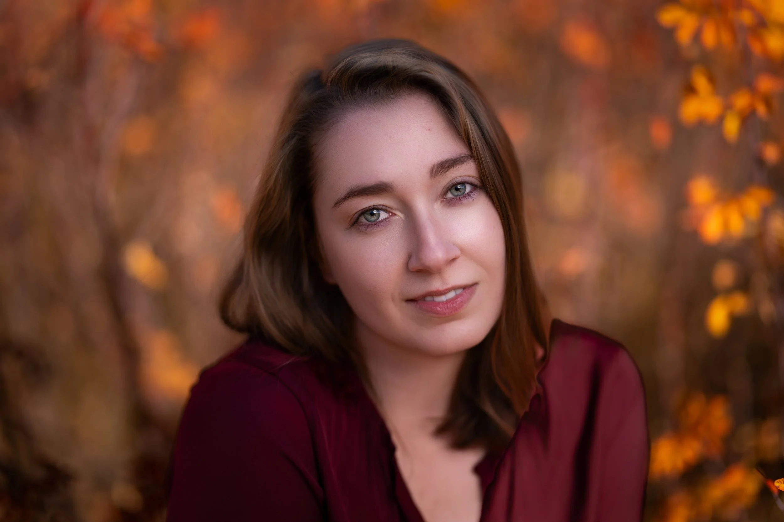 A woman with brown hair and blue eyes, wearing a maroon top, posing outdoors with autumn leaves in the background.
