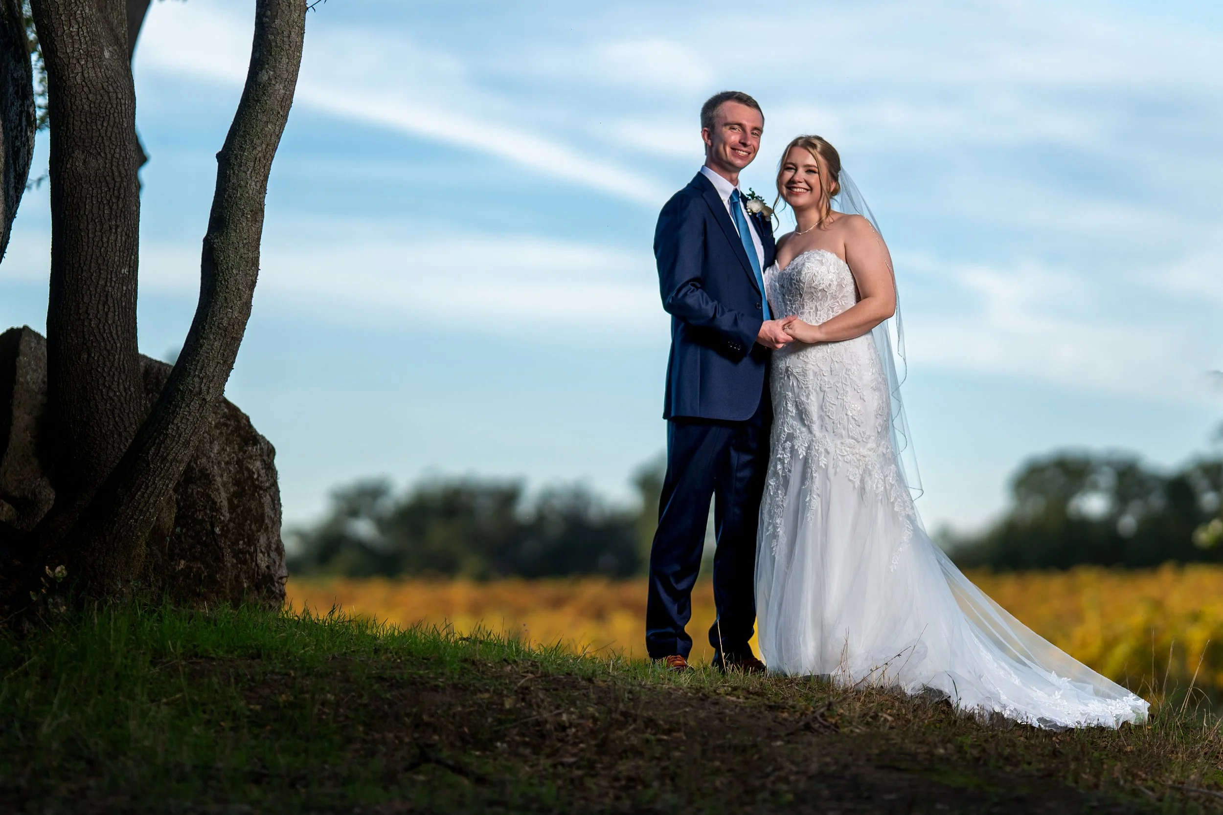 A newlywed couple dressed in wedding attire outdoors, standing on grass near trees, smiling at the camera with a cloudy sky background.
