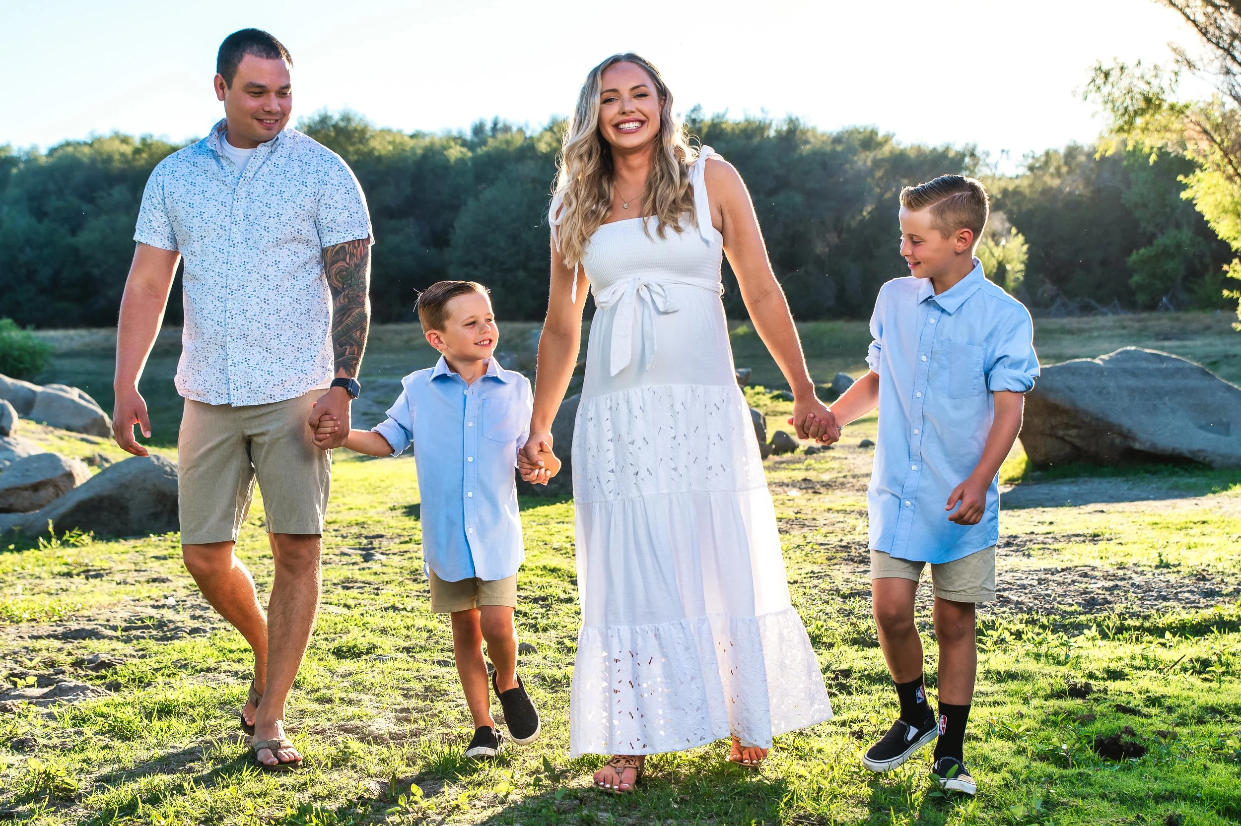 A family of five holding hands and walking outdoors in a park on a sunny day, with trees and rocks in the background. The woman is wearing a white dress and smiling at the camera.