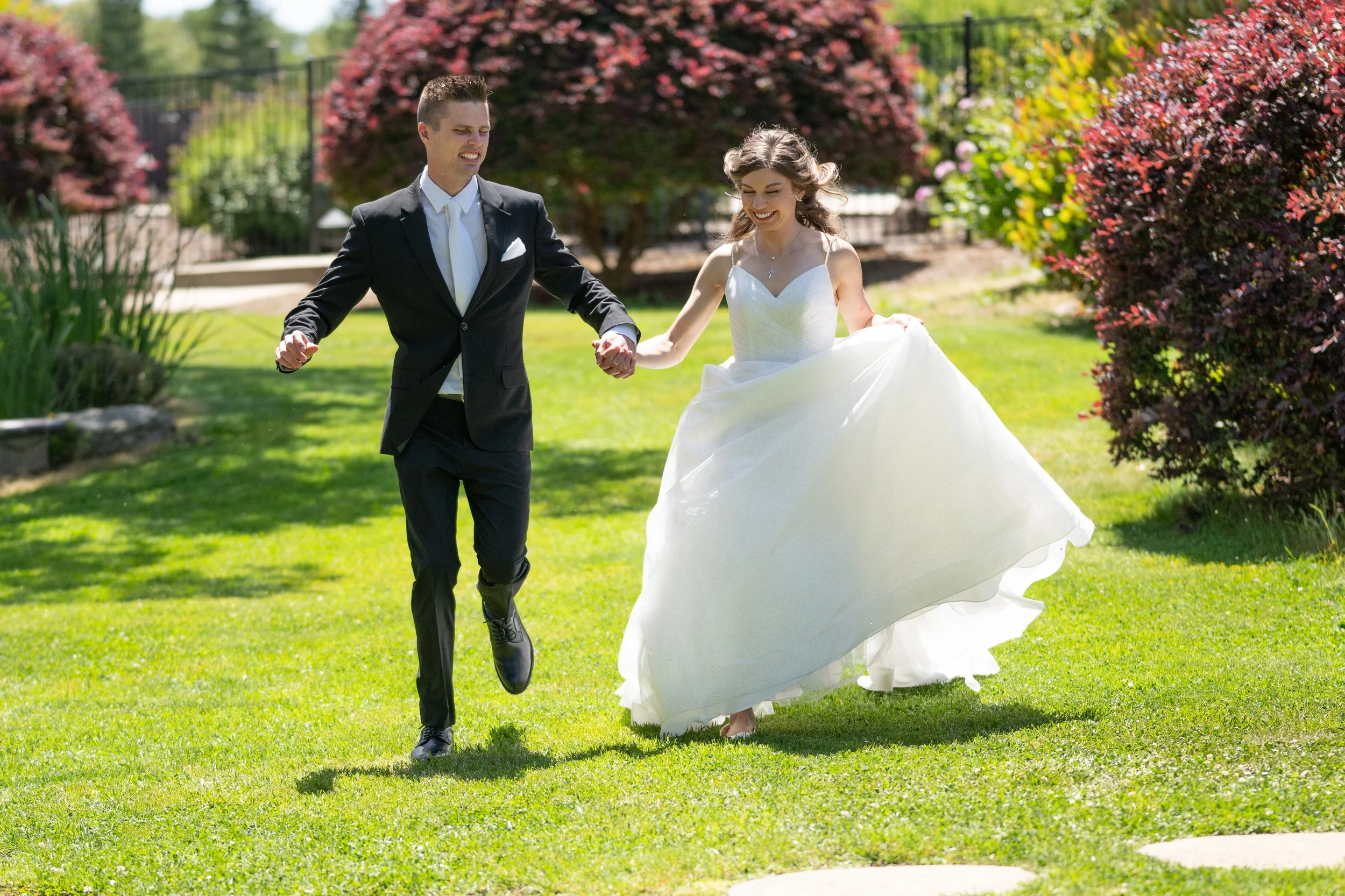A bride and groom holding hands and smiling while walking in a garden with colorful flowering bushes.