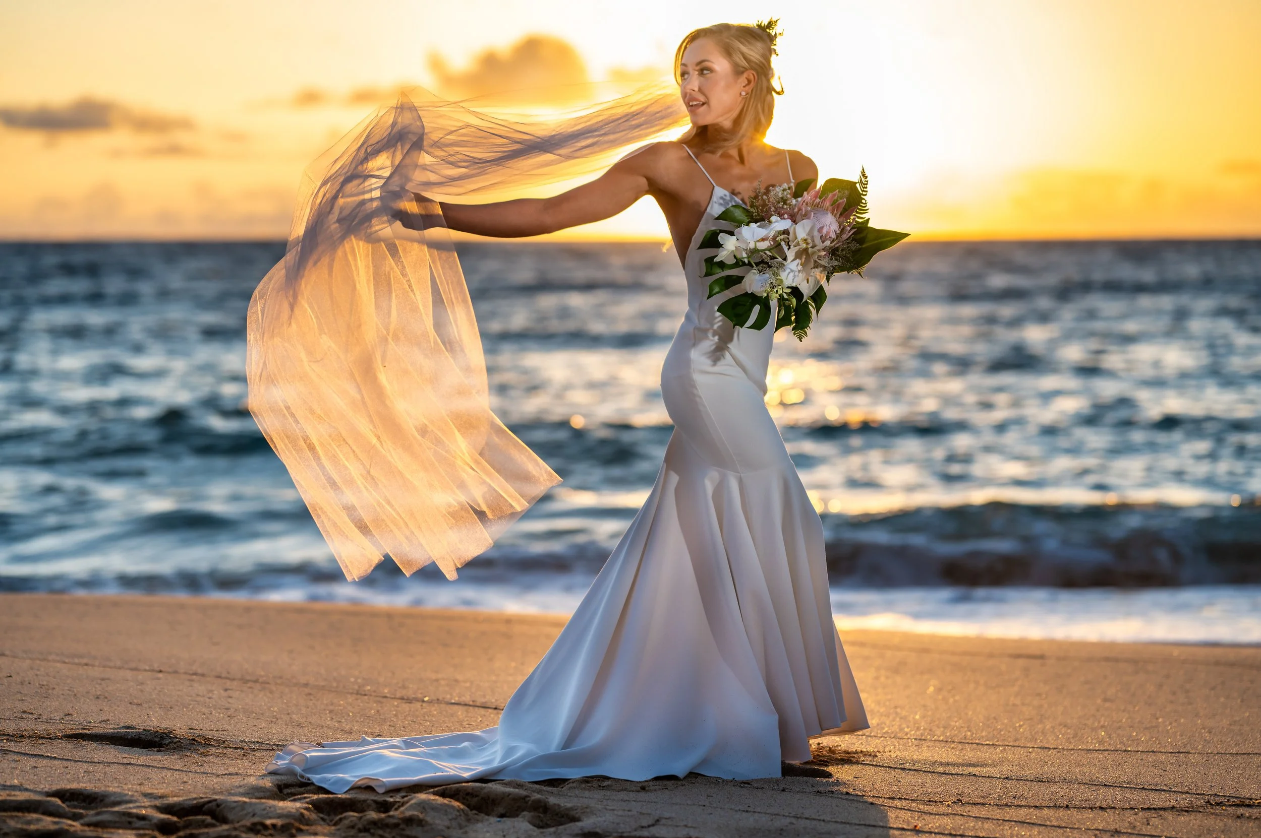 A woman in a white wedding dress holding a bouquet of flowers, standing on a beach at sunset.