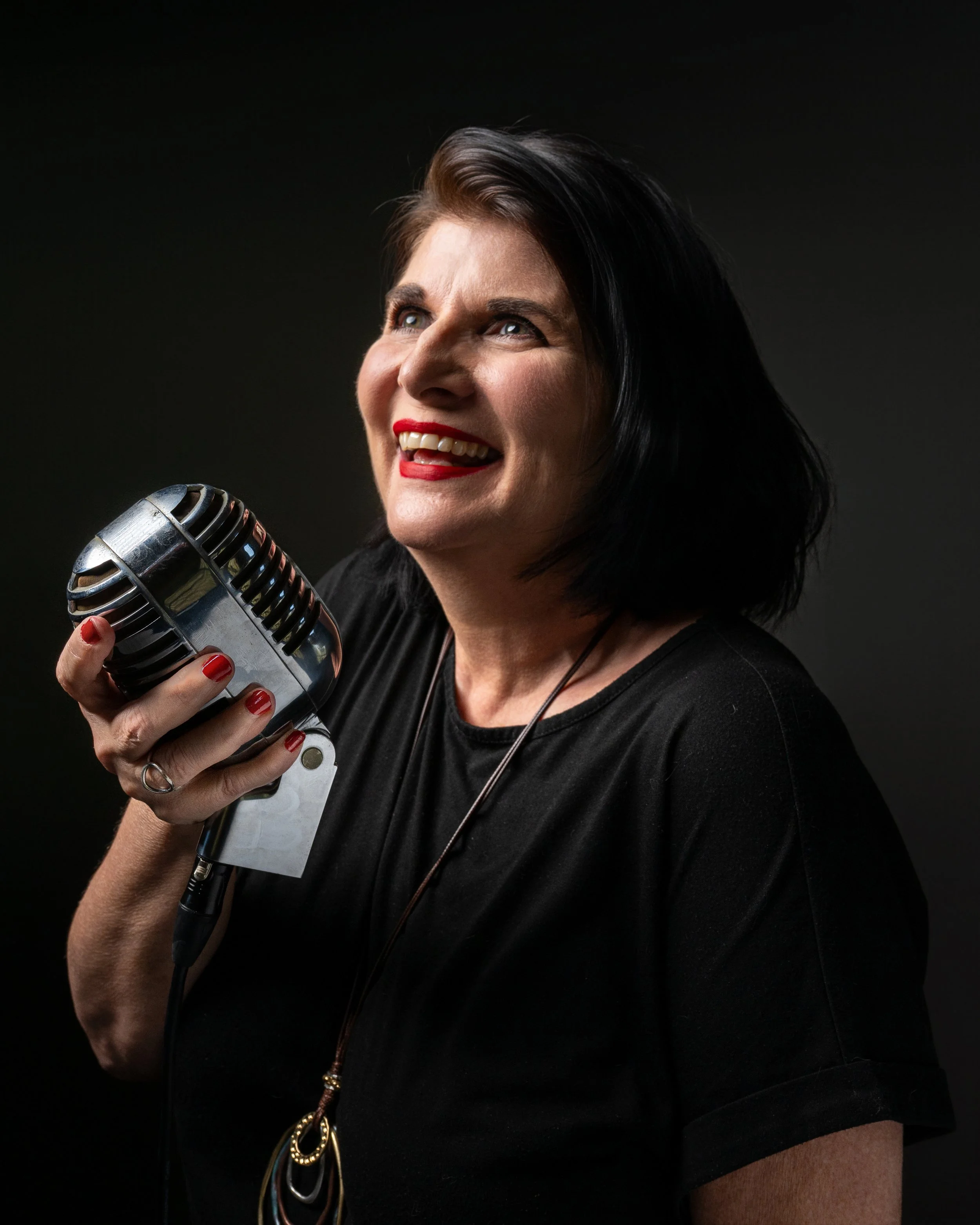 A woman with dark hair, red lipstick, and red nail polish holding a vintage microphone, smiling, against a black background.