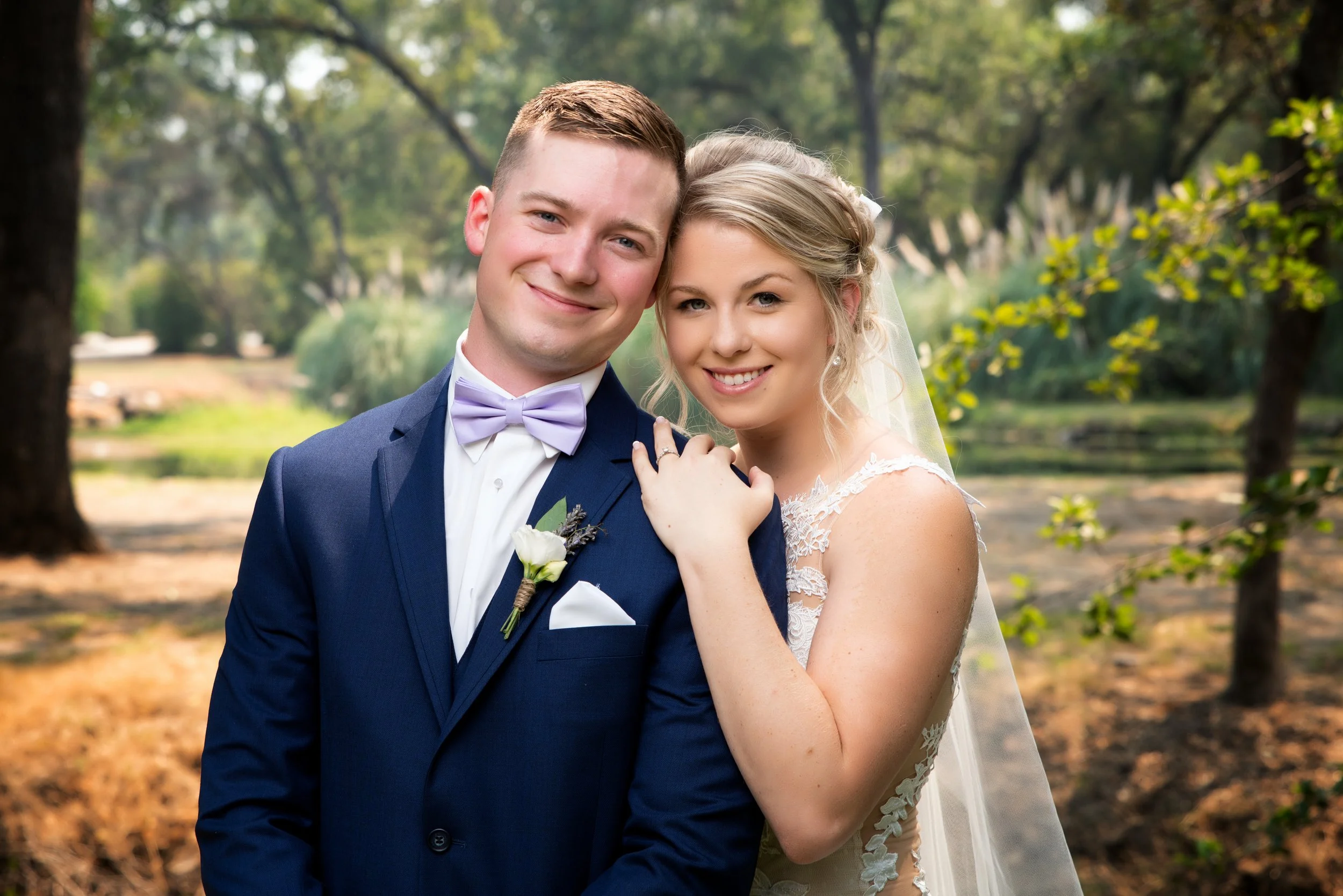 A young couple dressed in wedding attire, standing outdoors in a park or garden, smiling and hugging on a sunny day.