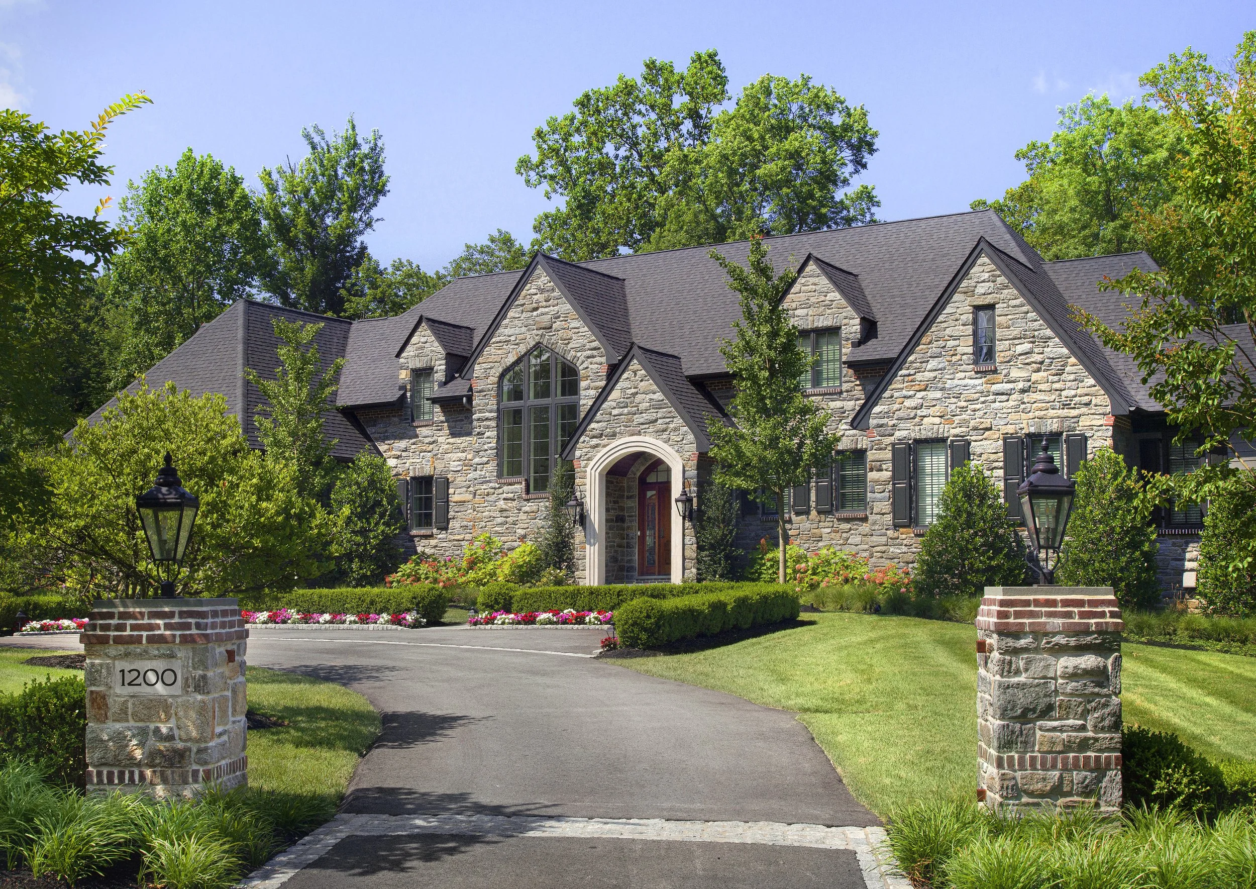 Large stone house with black roof, arched doorway, and multiple windows surrounded by lush green trees and landscaping. Brick pillars with lamps line the driveway.