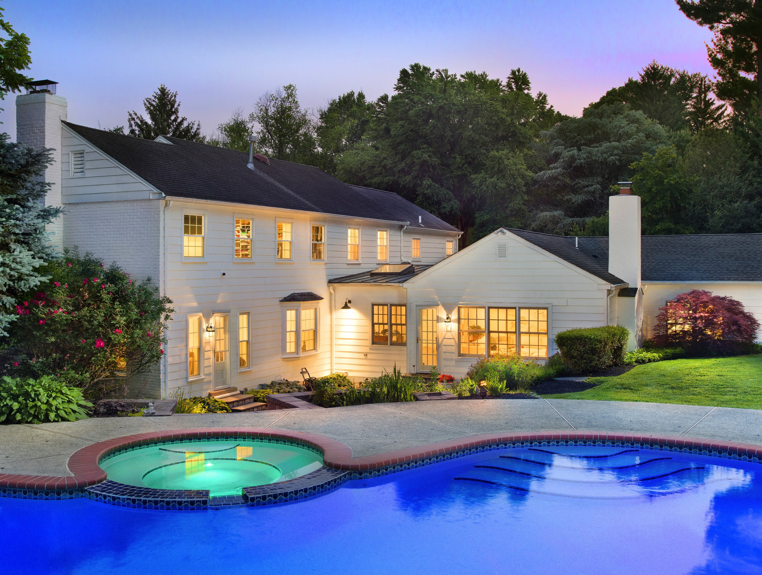 A backyard night scene showing a white house with lit windows, surrounded by lush green trees and bushes. In the foreground, a swimming pool and a hot tub are illuminated, creating a peaceful and inviting atmosphere.
