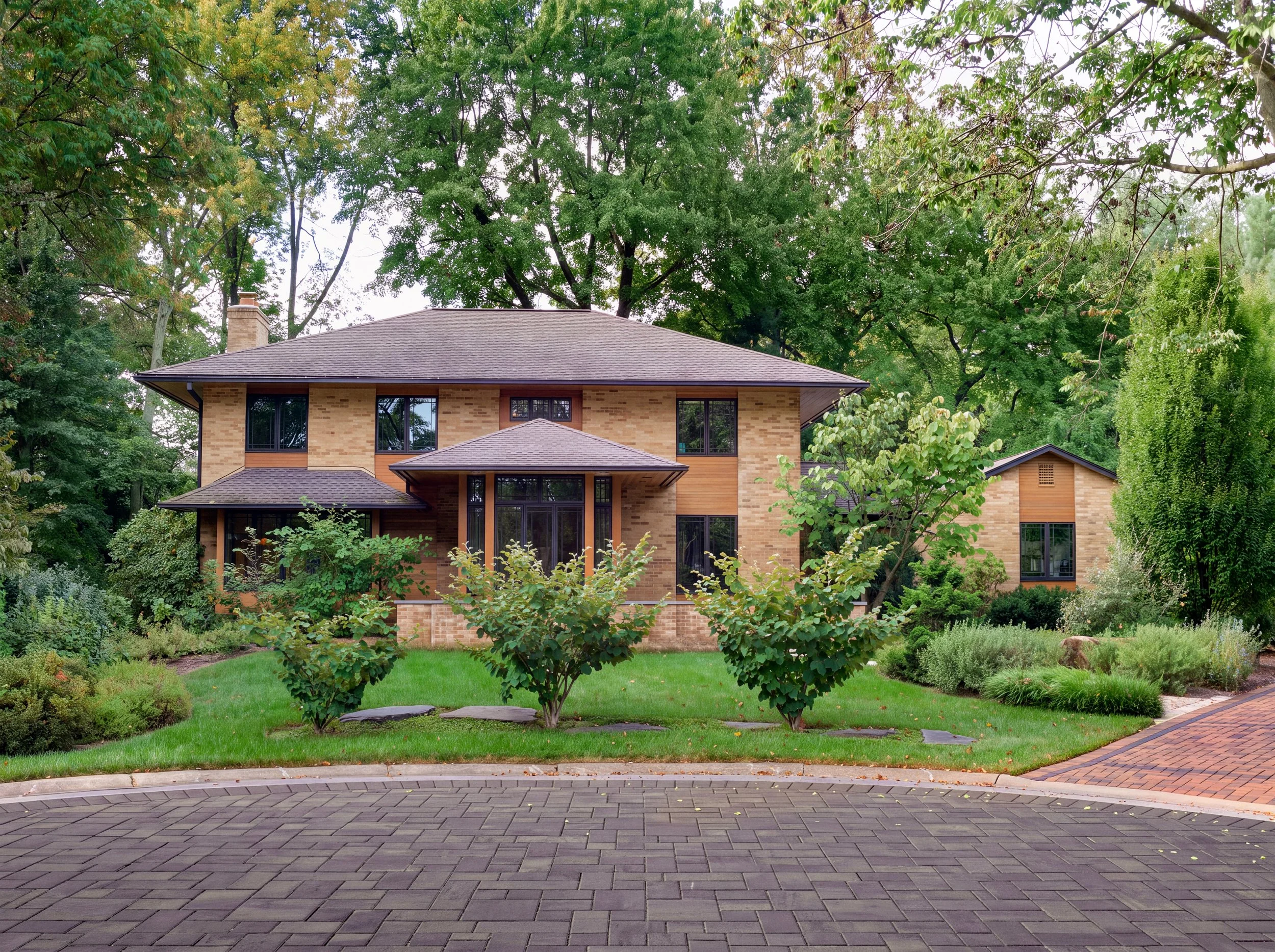 Large modern brick house with dark hipped roof, landscaped front yard, mature trees, and paved driveway