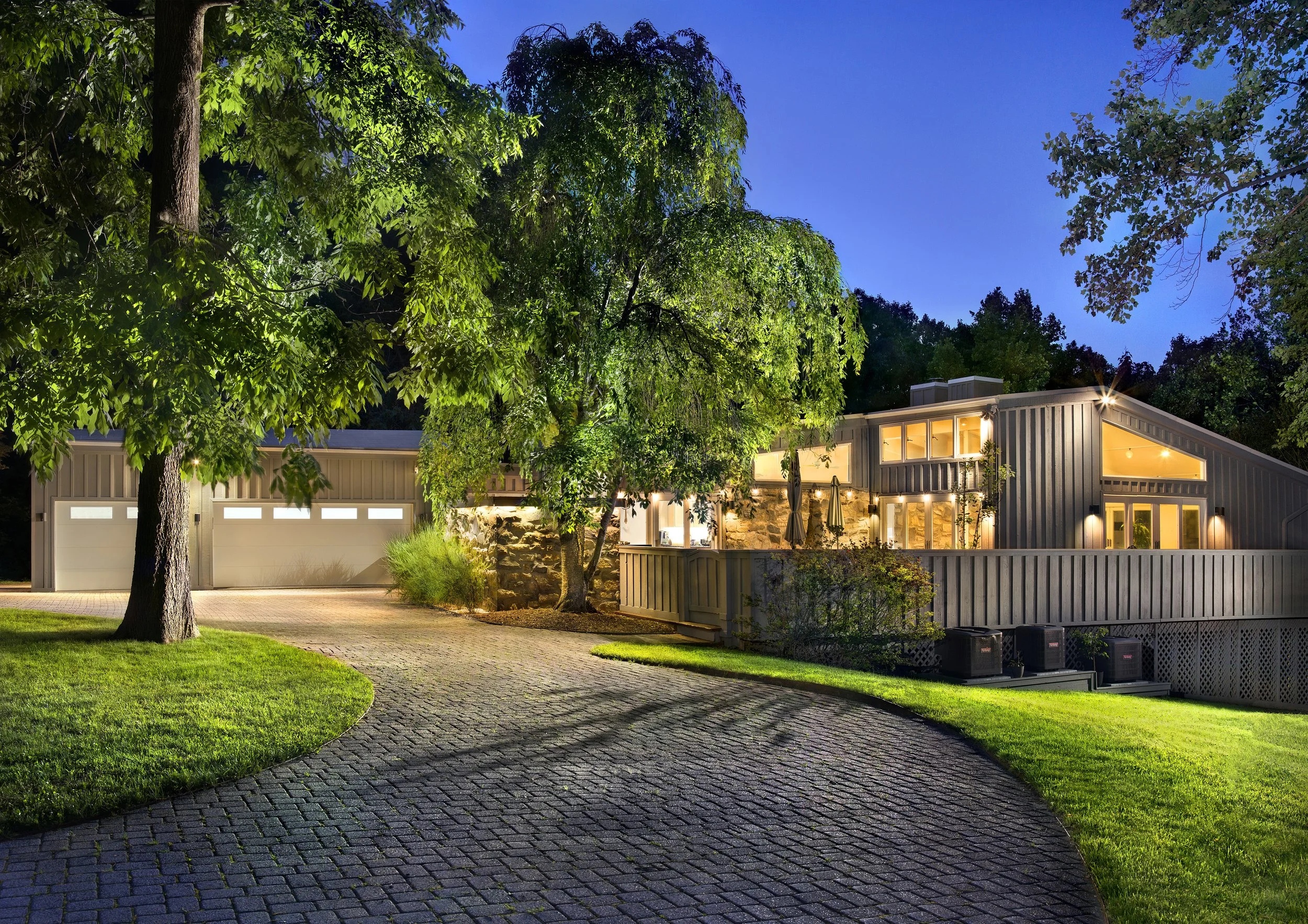 A modern house at dusk with large windows and warm interior lighting. The house is surrounded by a well-maintained green lawn, tall trees, and a cobblestone driveway. Outdoor lighting highlights the house and trees, creating a cozy atmosphere.