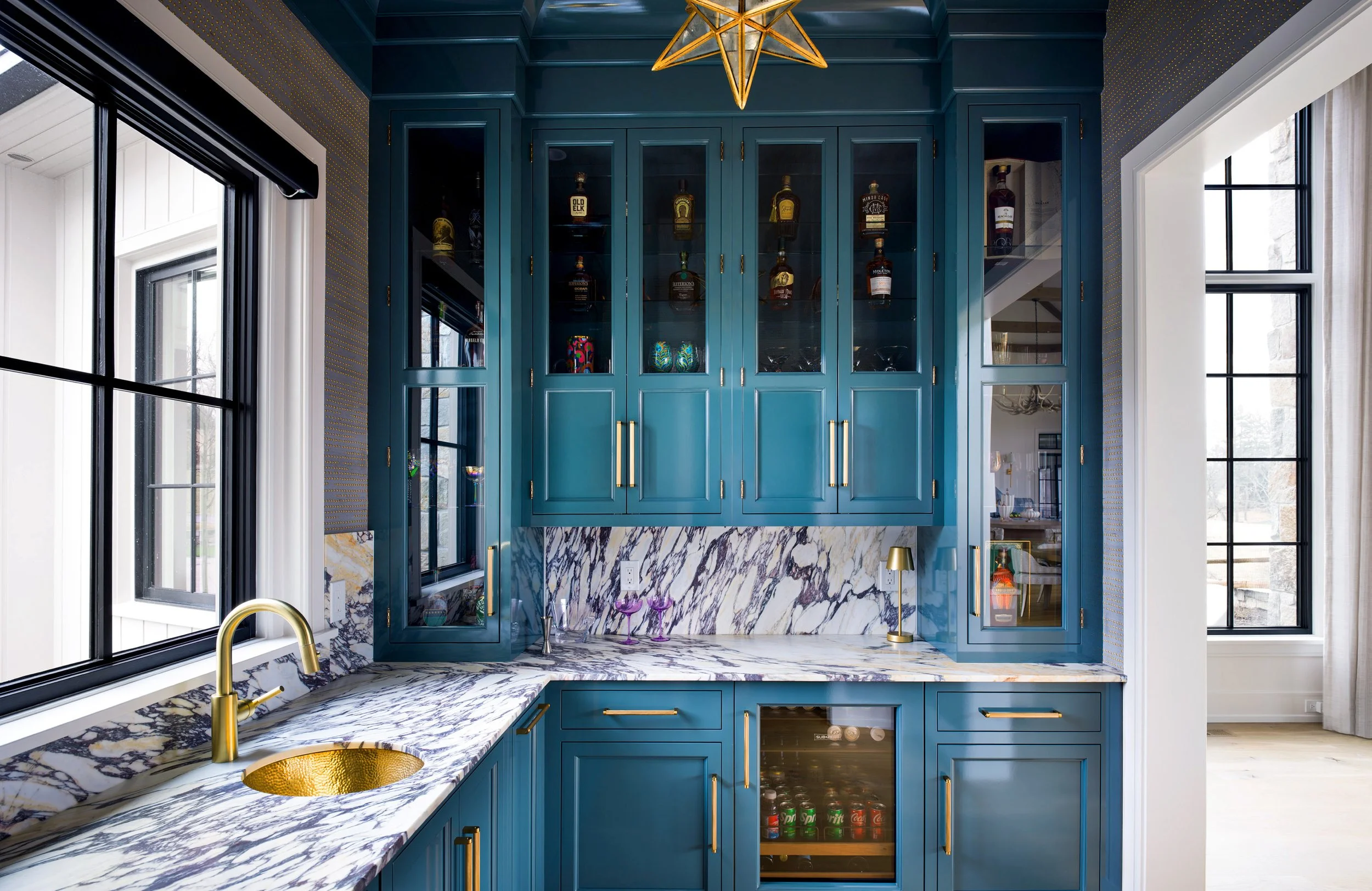 View of a blue kitchen with glass-front cabinets, marble countertops, a gold sink, and windows providing natural light.