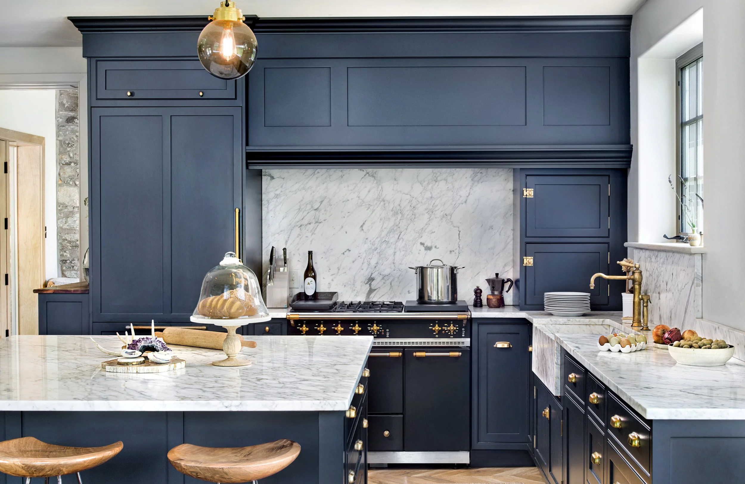 Modern kitchen with navy blue cabinets, white marble countertops, and a marble backsplash. Gold accents on hardware and fixtures, and a large island with a marble top and wooden stools. Items on countertops include a cake stand with cake under glass,