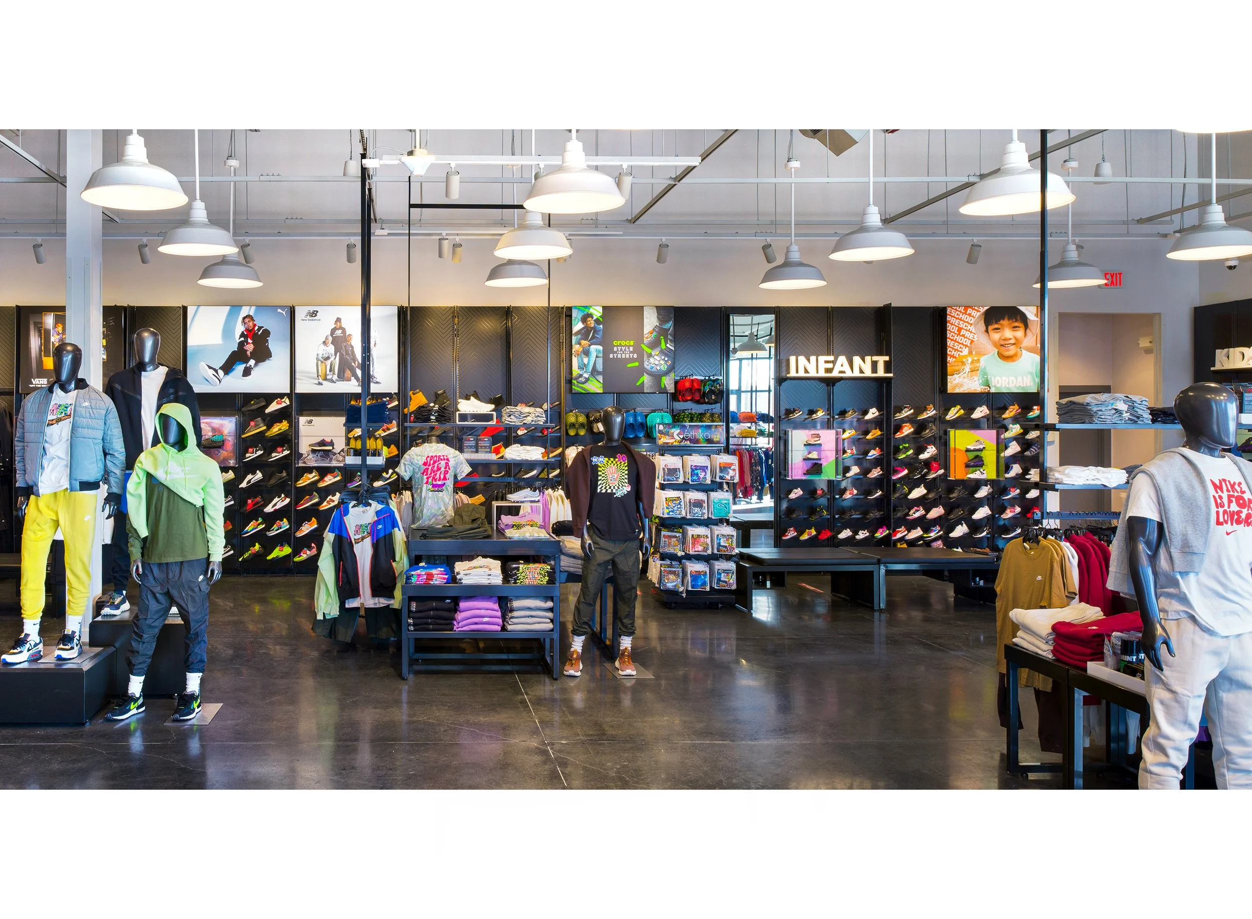 Inside a retail store showing children's and athletic clothing and shoes, with racks of clothes, mannequins, and shelves filled with shoes, displayed under bright white pendant lighting. Athletic