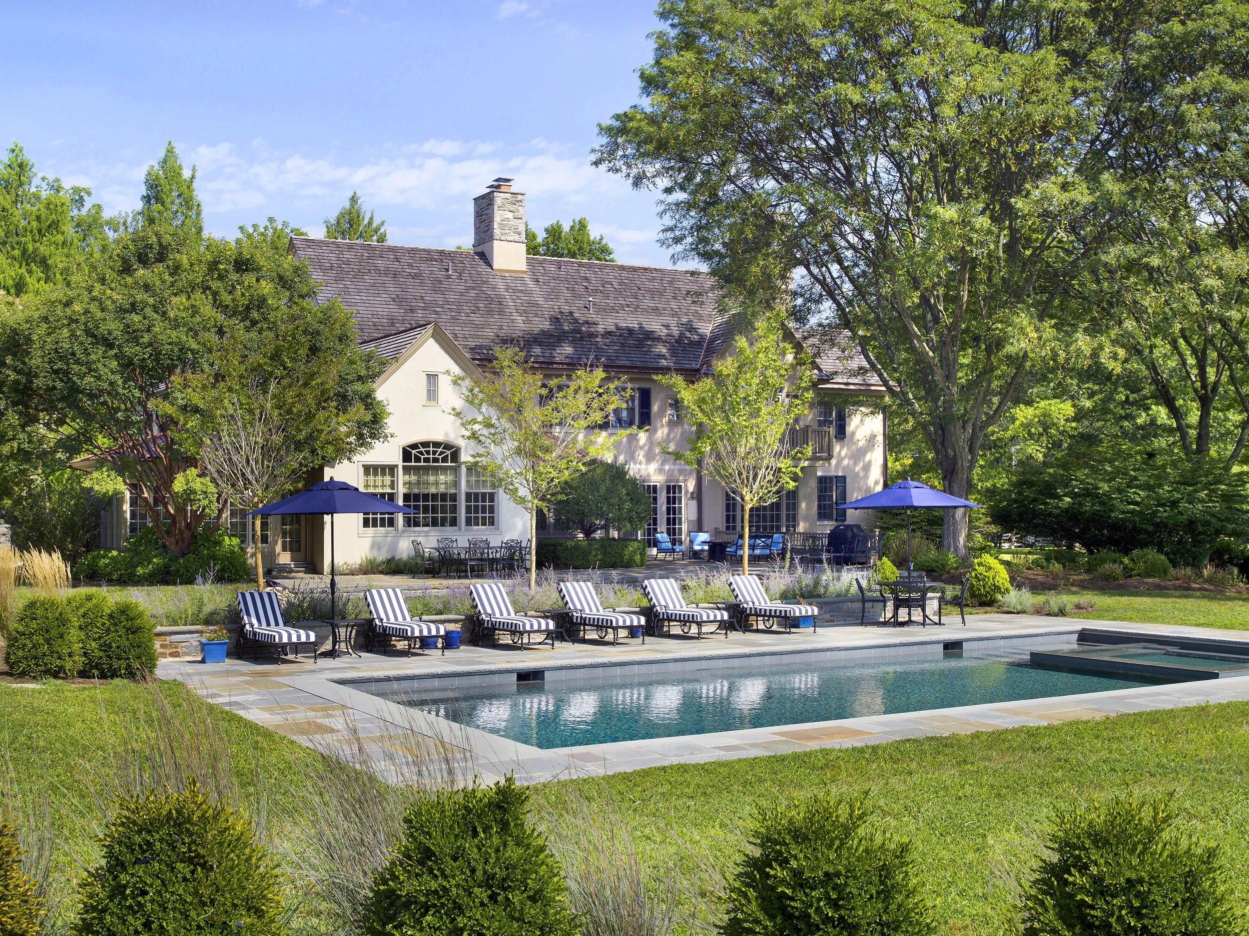 Backyard view of a house with a swimming pool in the foreground, surrounded by greenery, trees, and outdoor furniture with umbrellas under a blue sky.