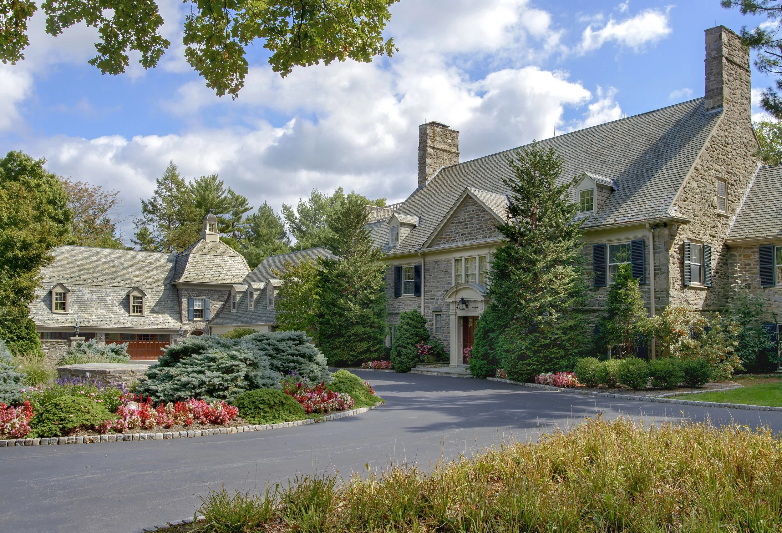 A large stone house with multiple chimneys, surrounded by well-maintained garden and tall trees, under a partly cloudy sky.