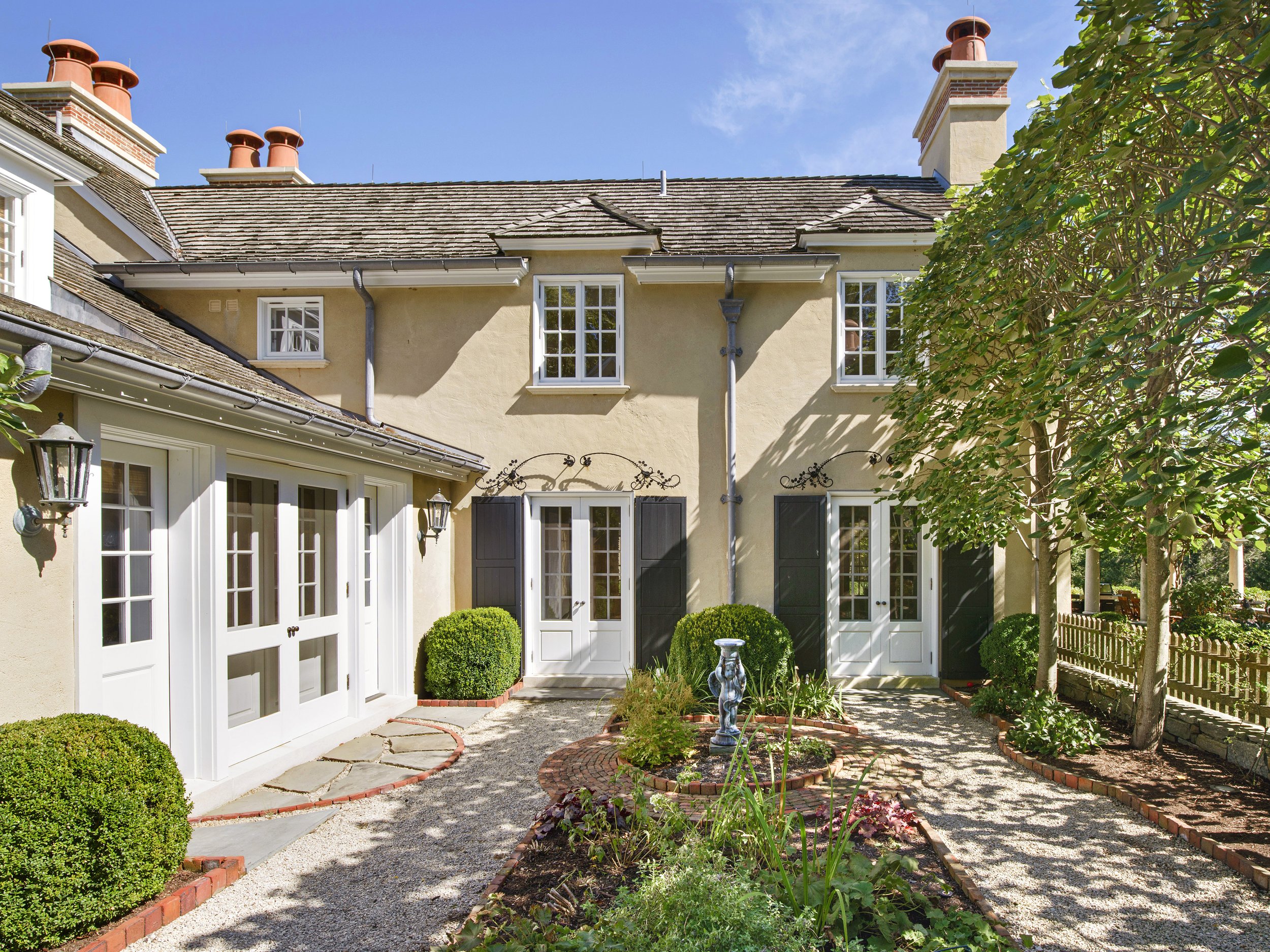A backyard view of a yellow house with large white-framed windows, dark shutters, and multiple chimneys on the roof. The yard features a gravel pathway, well-maintained bushes, a small garden with plants, and a decorative garden sculpture. There is a