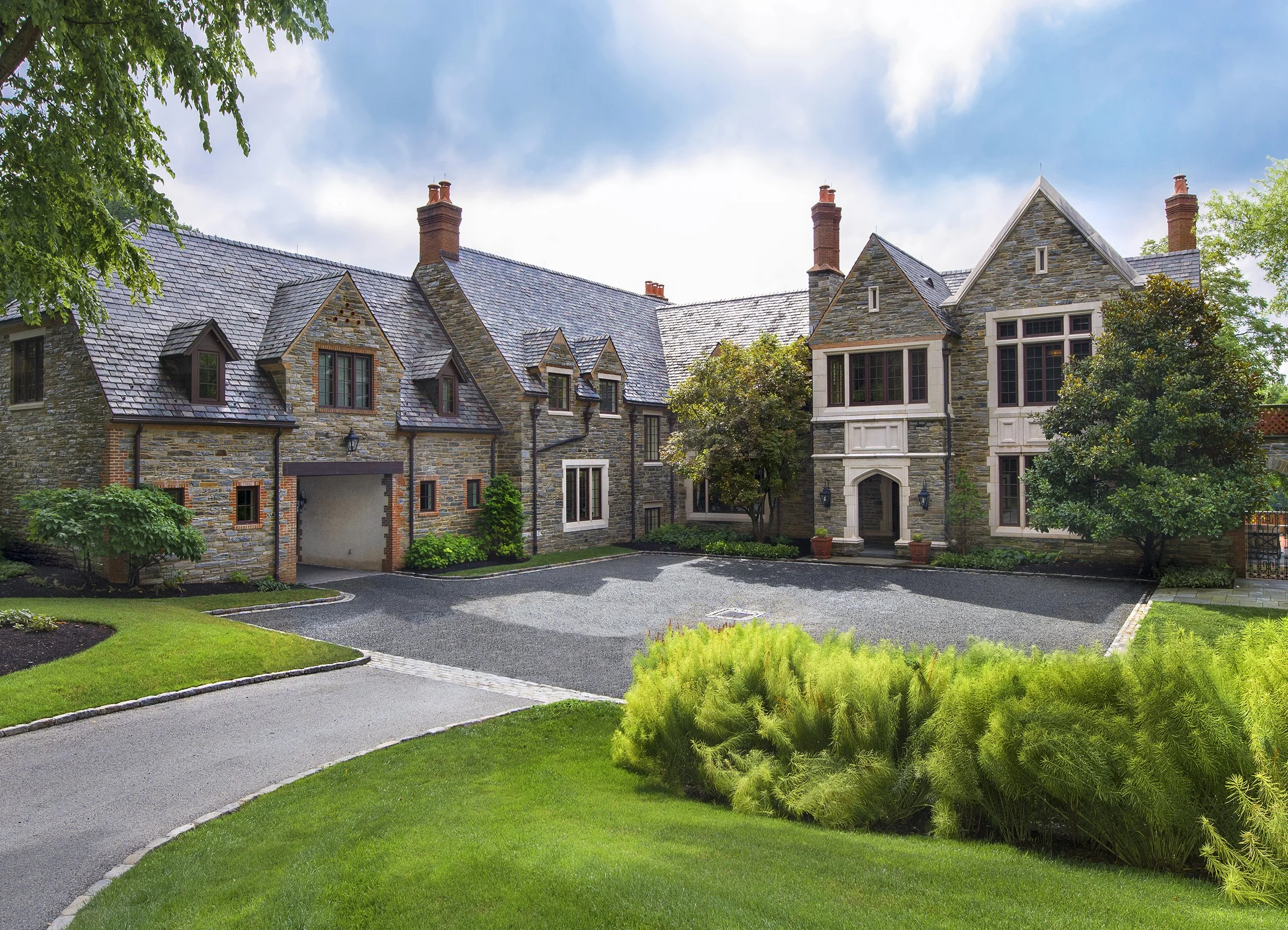 A large, elegant stone house with multiple gabled roofs topped with slate tiles, tall brick chimneys, and ornate windows, surrounded by a lush, landscaped garden with neatly trimmed grass, various trees, and a paved driveway.