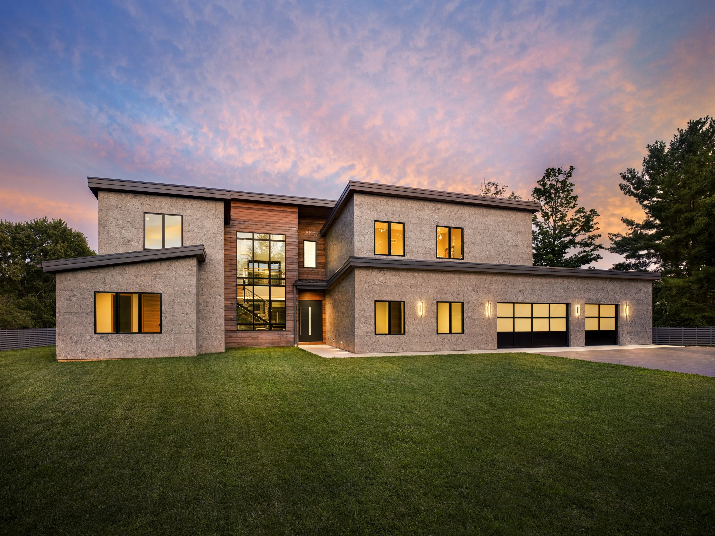 Modern multi-story house with large windows and a flat roof, surrounded by green lawn and trees at sunset, with a colorful sky in the background.