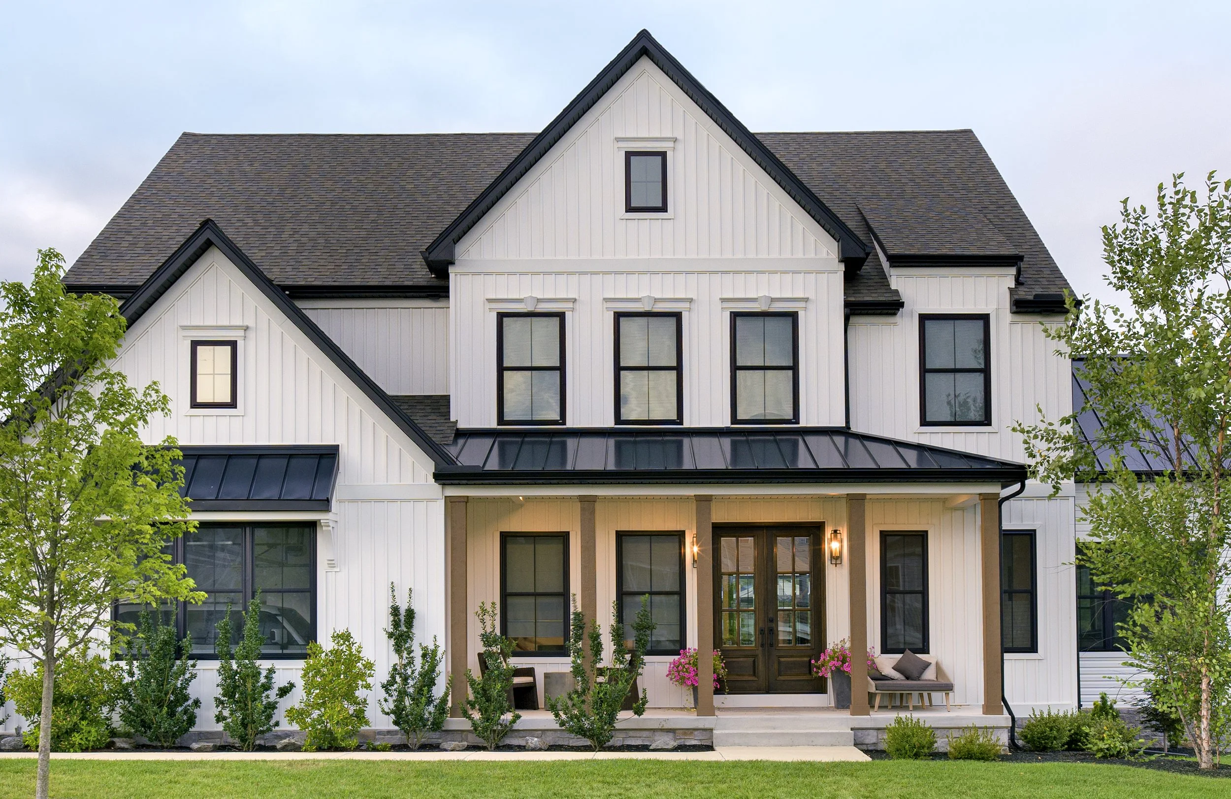 Front view of a modern farmhouse-style house with white vertical siding, black window frames, a dark roof, and a front porch with seating and blossoming pink plants.