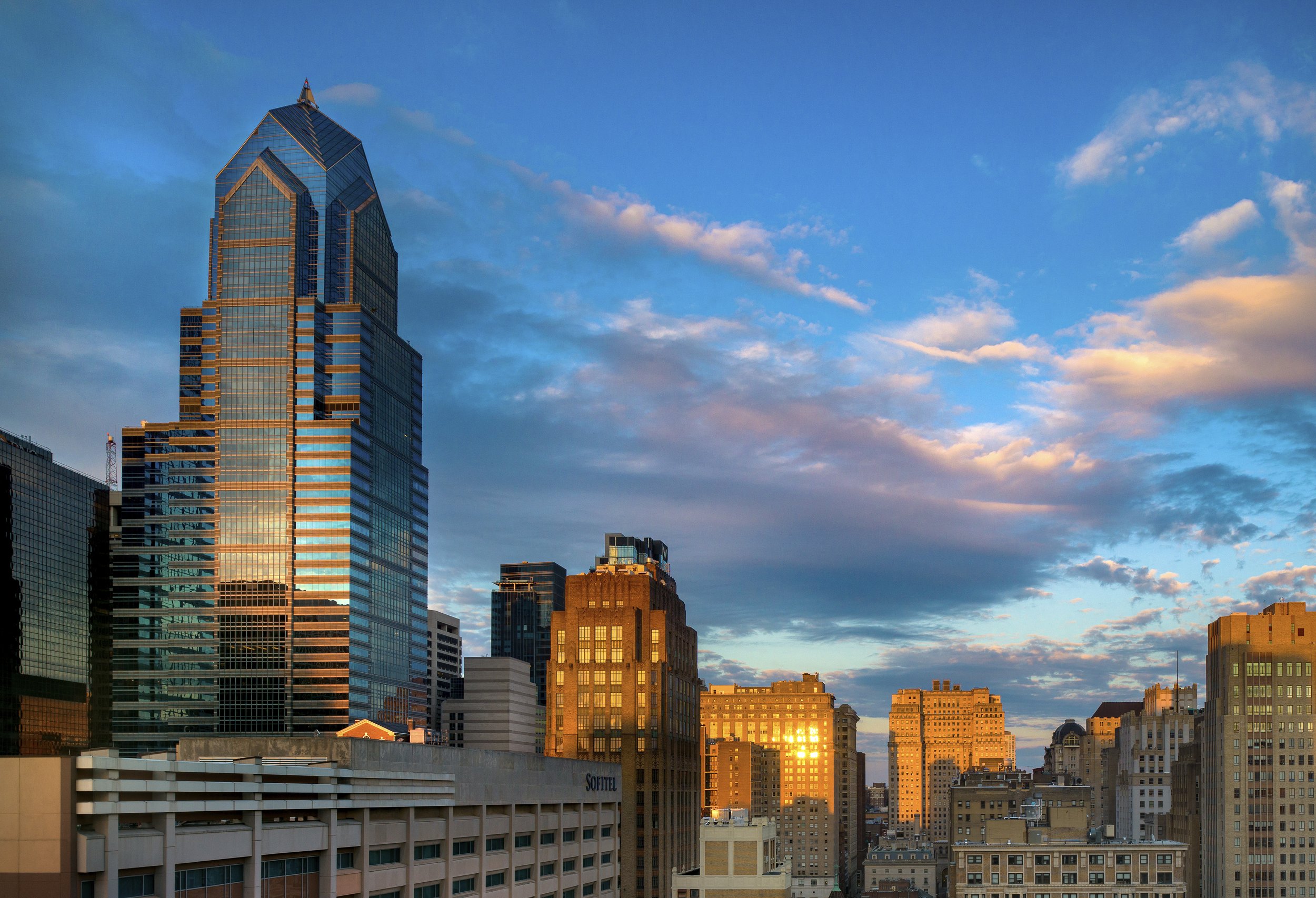 Skyline of a city with modern and older buildings at sunset, with the sun reflecting off windows and a blue sky with scattered clouds.