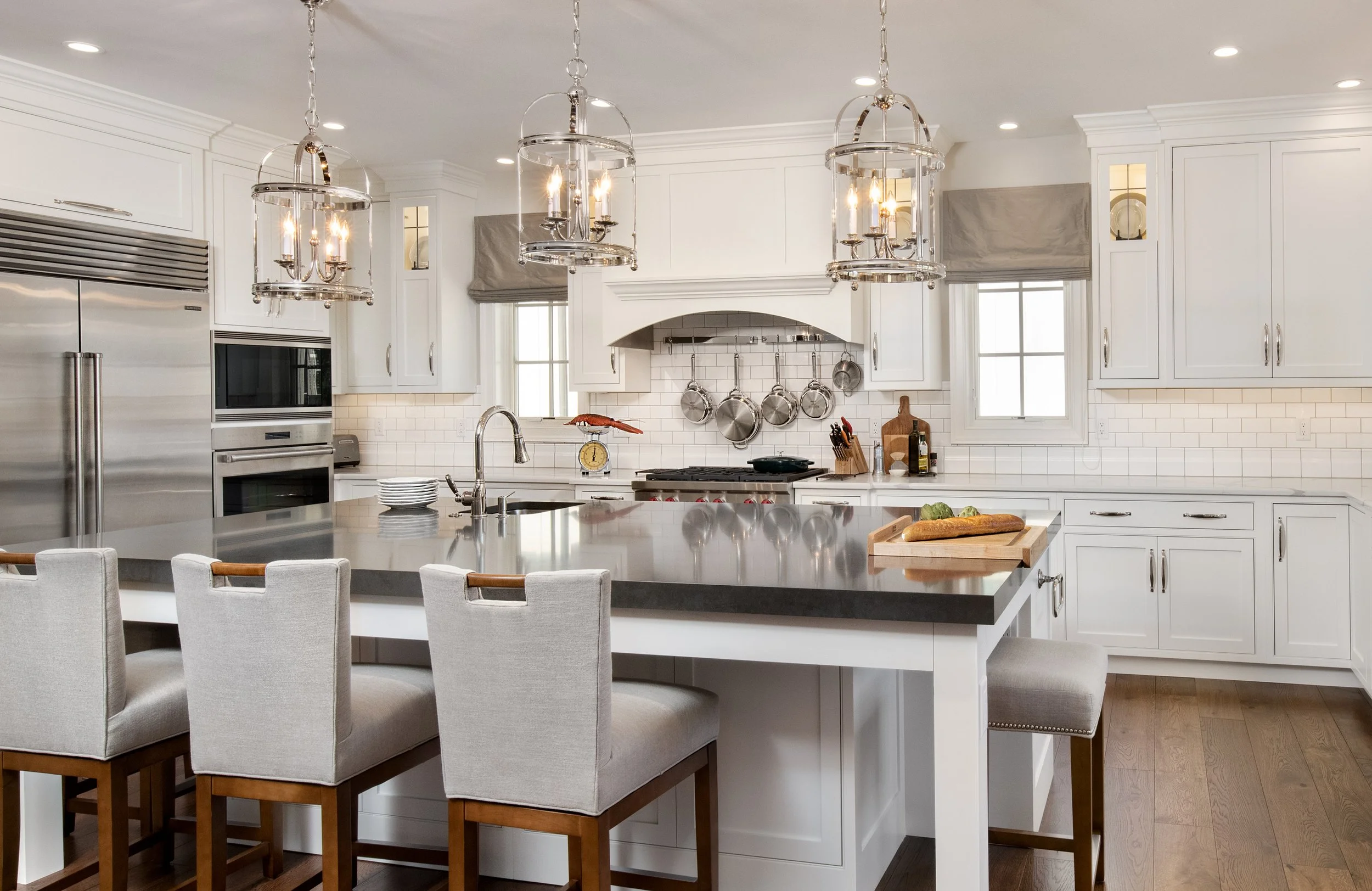 Modern white kitchen with a large island, stainless steel appliances, three hanging chandelier lights, and a row of four upholstered chairs at the island.