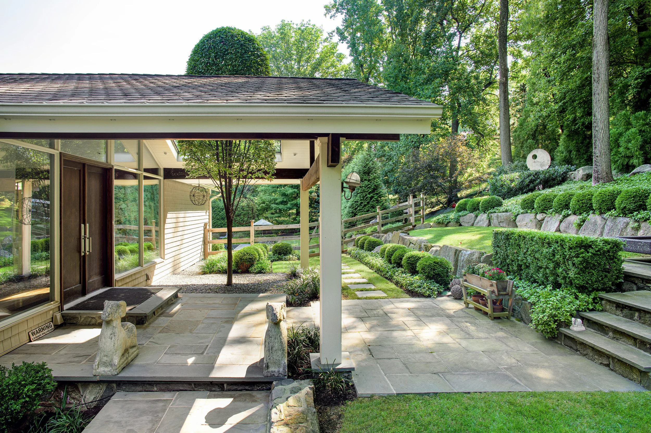 Entryway patio with stone flooring, a small tree, stone dog sculptures, a landscaped garden with trimmed bushes, and steps leading to a grassy hill with large rocks and trees all done in mid-century style.