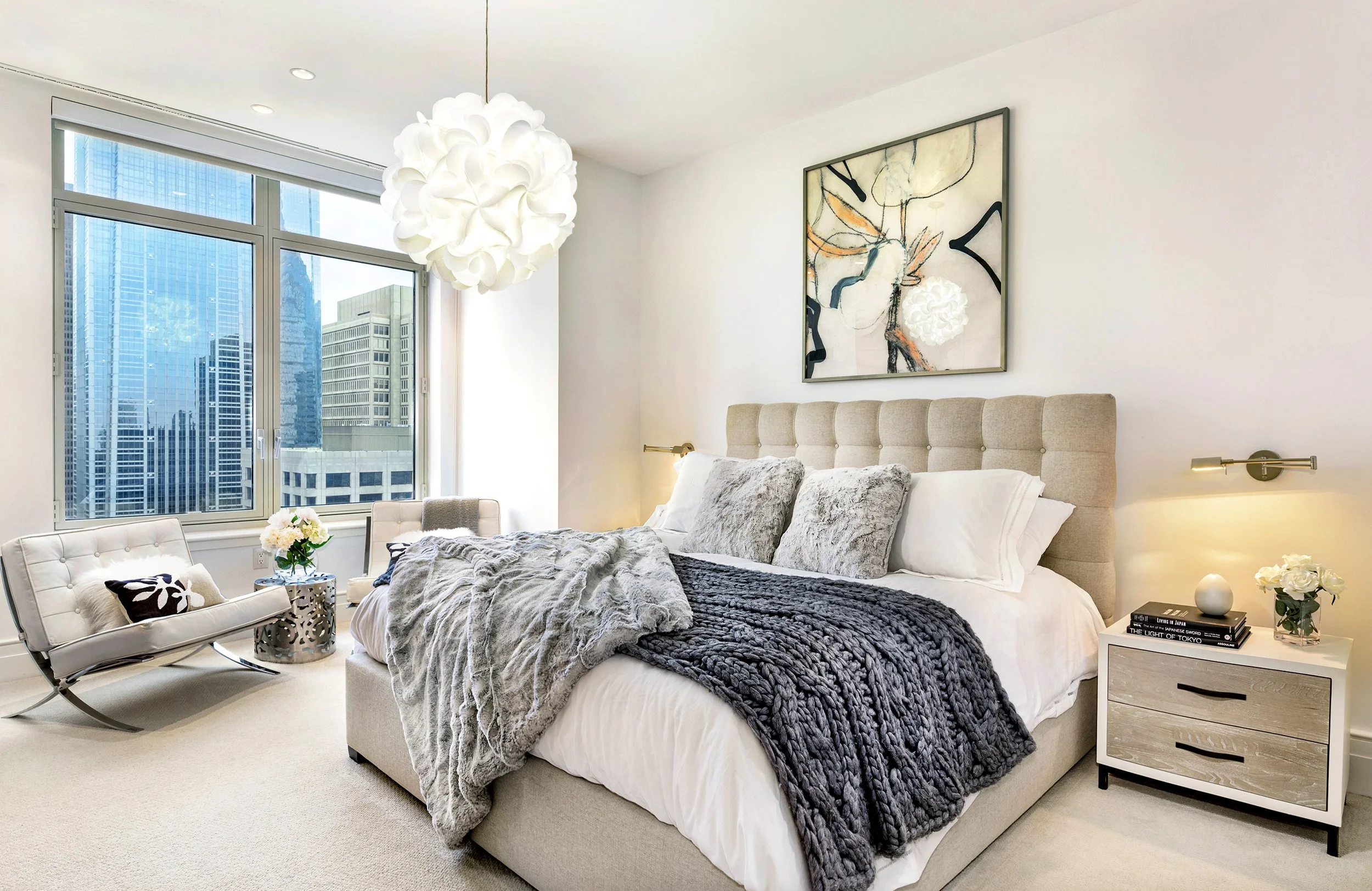 Modern bedroom with large window view of city skyscrapers, beige upholstered bed with plush pillows, abstract artwork on wall, white nightstand with flowers, and white lounge chair by window.