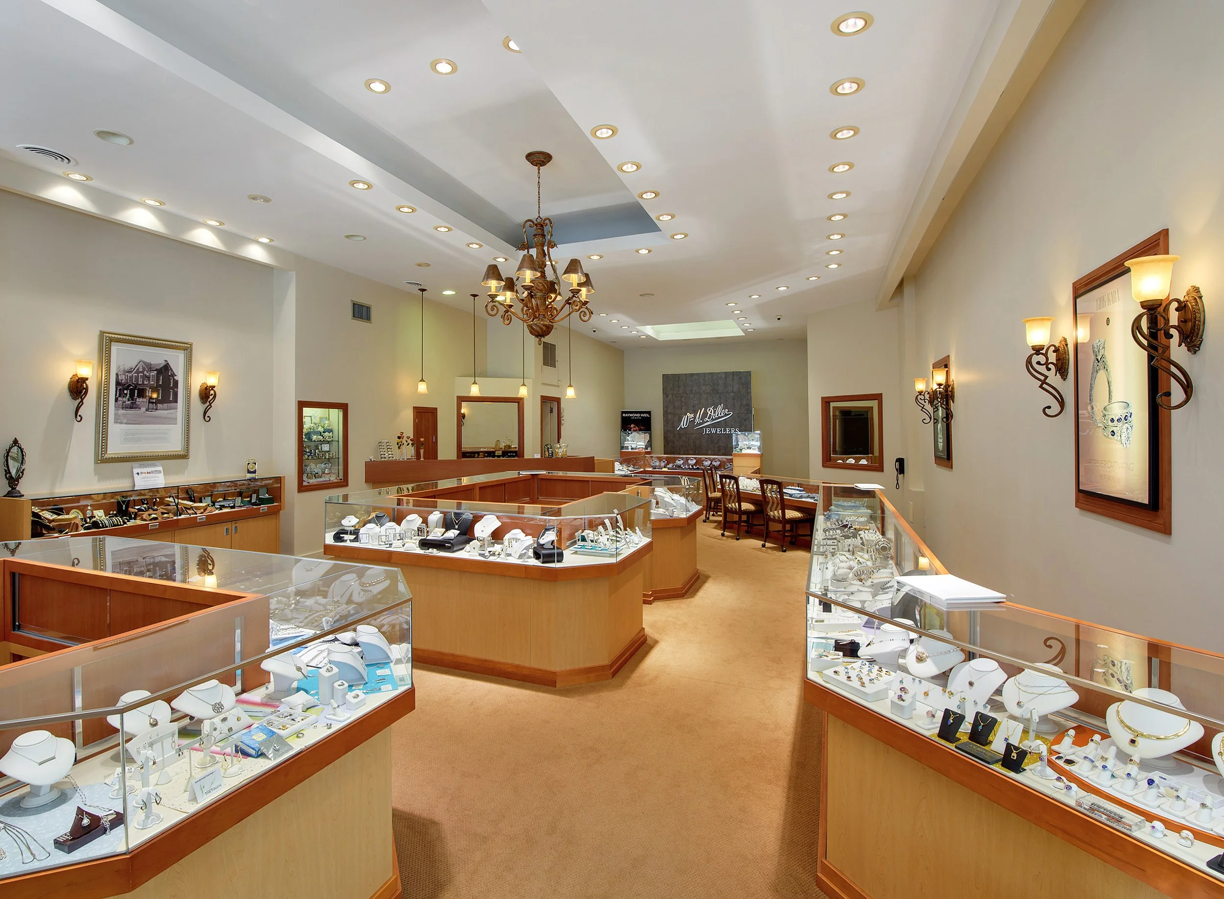 Interior of a jewelry store with display cases filled with necklaces, rings, and other jewelry, gold-colored lighting fixtures, and framed artwork on the walls.