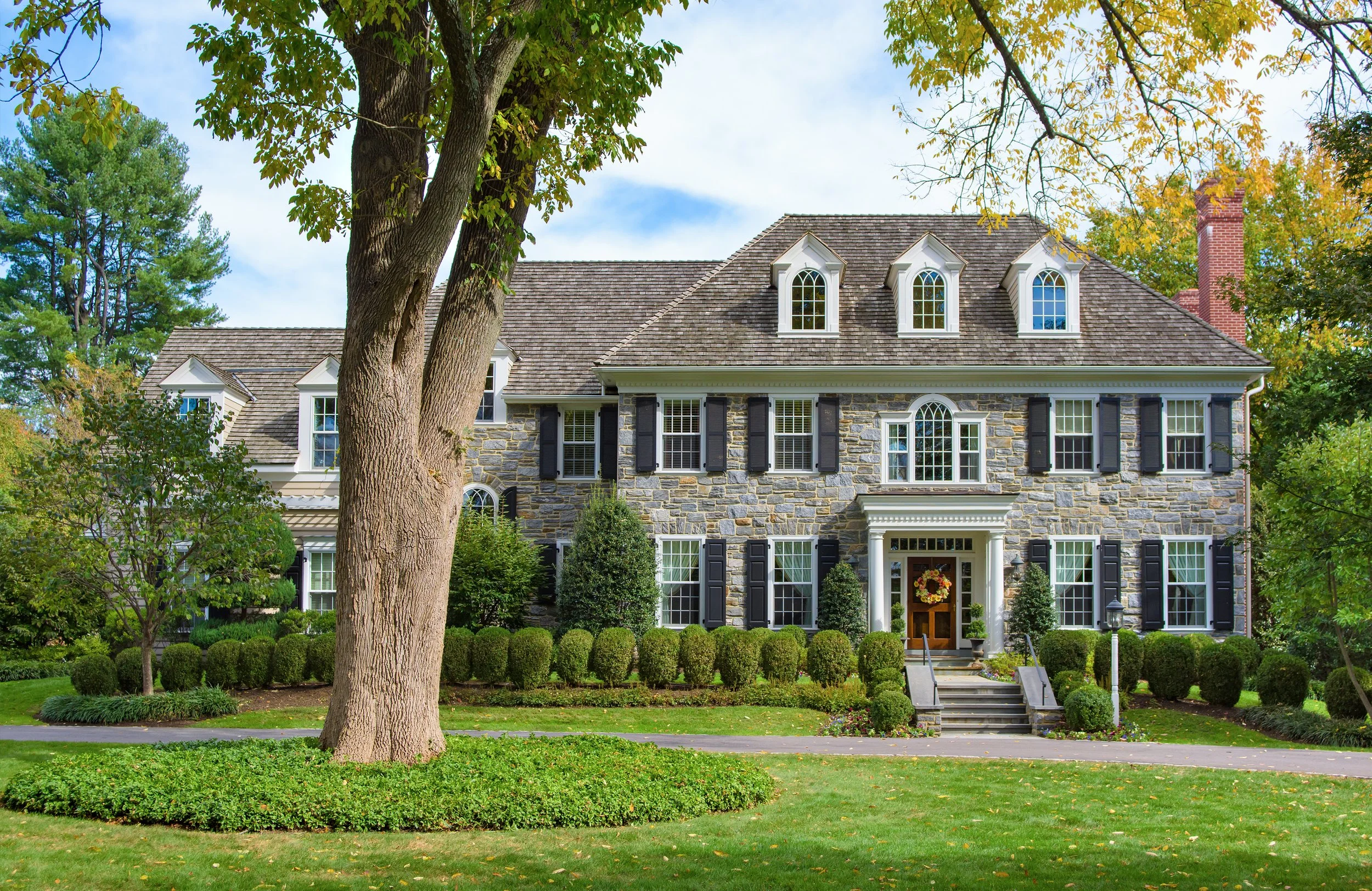 A large house with a stone facade, multiple dormer windows with arched tops, black shutters, and a front porch decorated with fall wreath. The house is surrounded by lush, well-maintained landscaping, including a large tree, shrubs, and a neatly trim