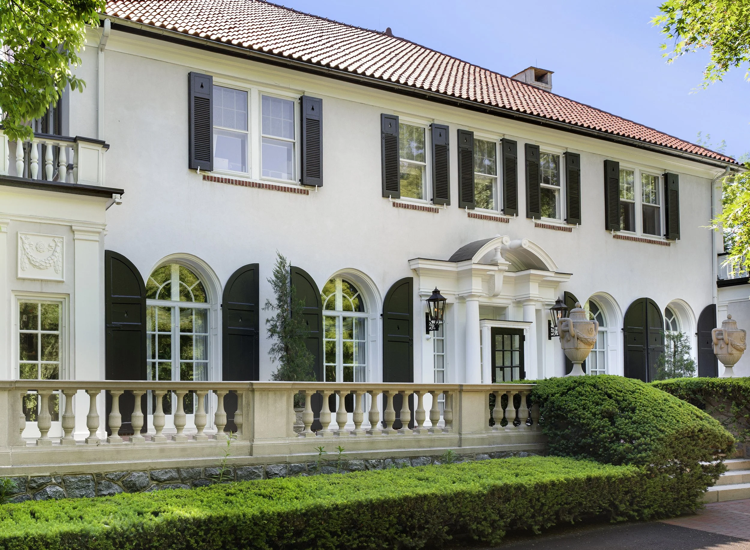 A large, elegant house with white exterior walls, black shutters, and a red-tiled roof. There is an ornate white railing on a balcony, well-maintained bushes, and trees around the house. The windows are tall with decorative arches.