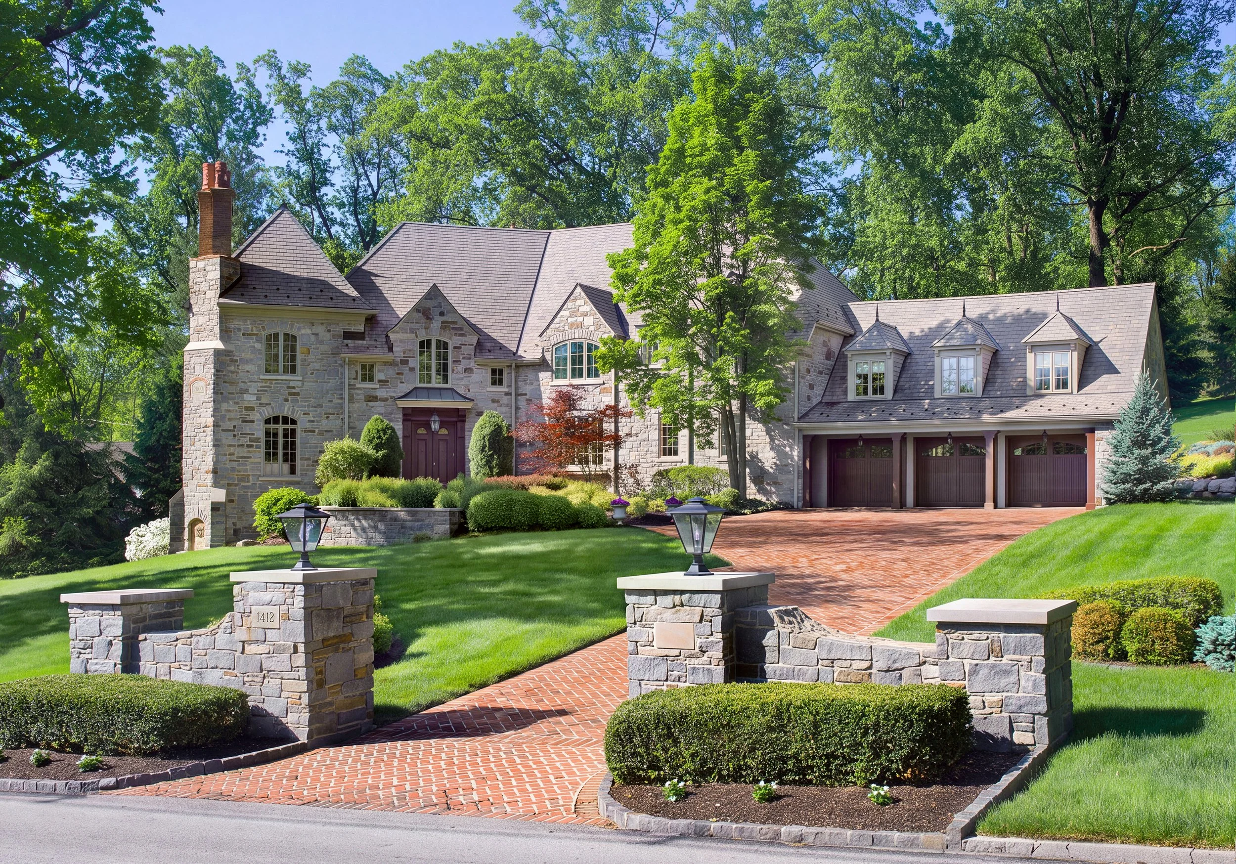 Large stone luxury home with multiple gables, brick driveway, manicured landscaping, and mature trees