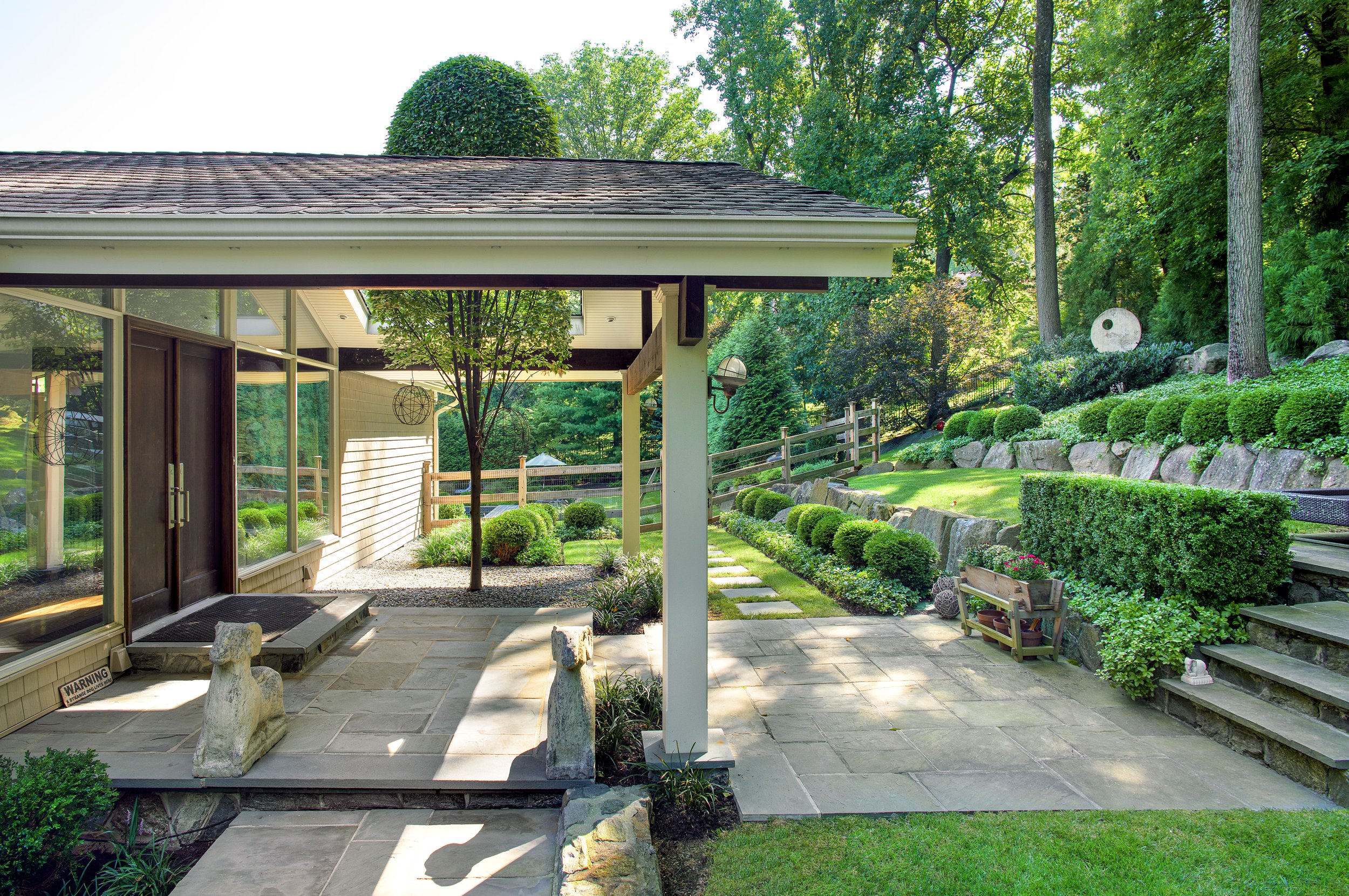Backyard patio with stone flooring, a small tree, stone dog sculptures, a landscaped garden with trimmed bushes, and steps leading to a grassy hill with large rocks and trees.